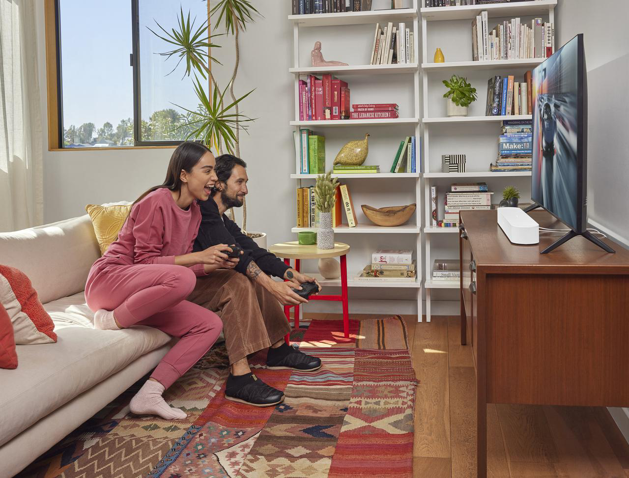 Ein Paar spielt Videospiele auf einem Sofa. Rechts ist ein Fernseher, der das Spiel zeigt.