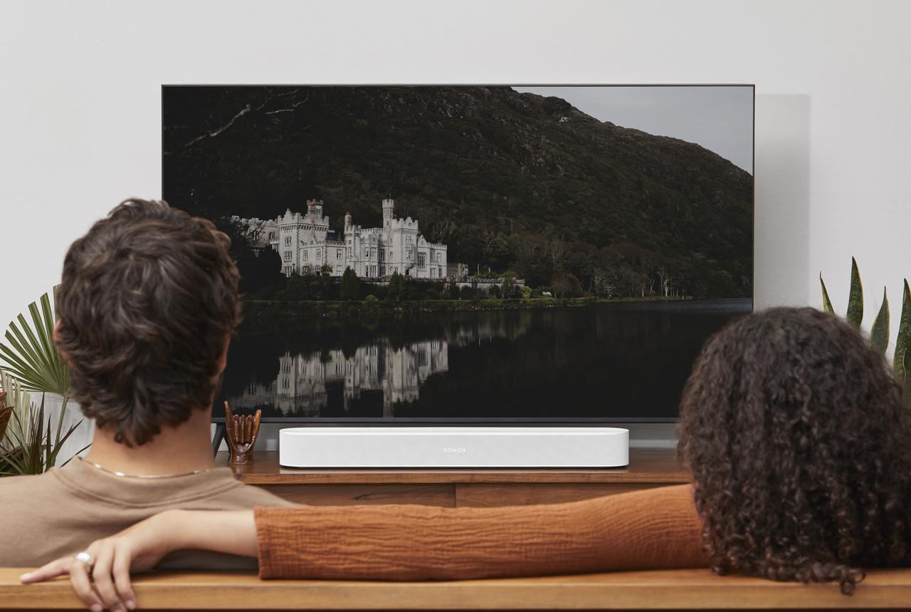 Ein Paar sitzt auf einer Holzbank und schaut einen Fernseher mit einer Sonos Soundbar.