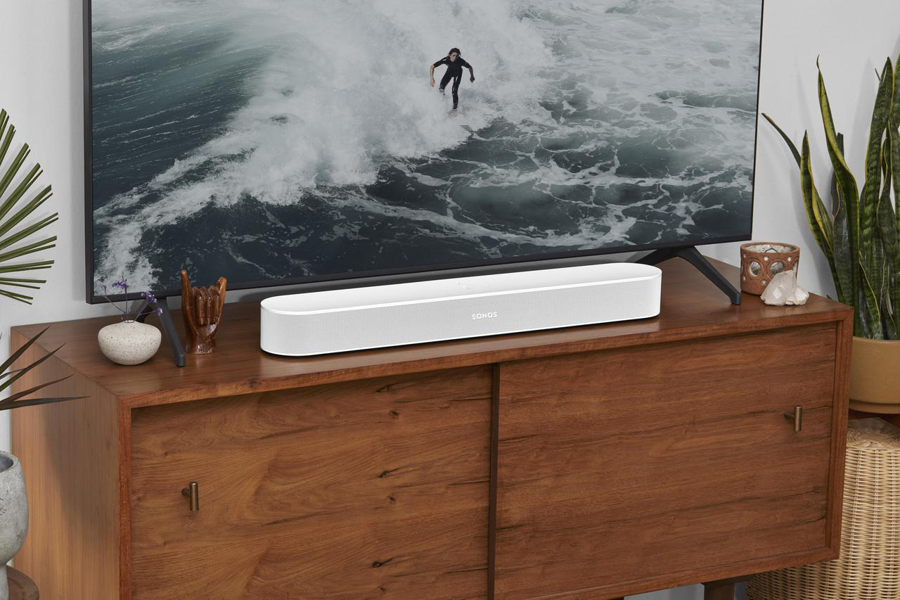 Eine weiße Soundbar auf einem Holzschrank, mit einem Fernseher, der einen Surfer zeigt, und Pflanzen im Hintergrund.