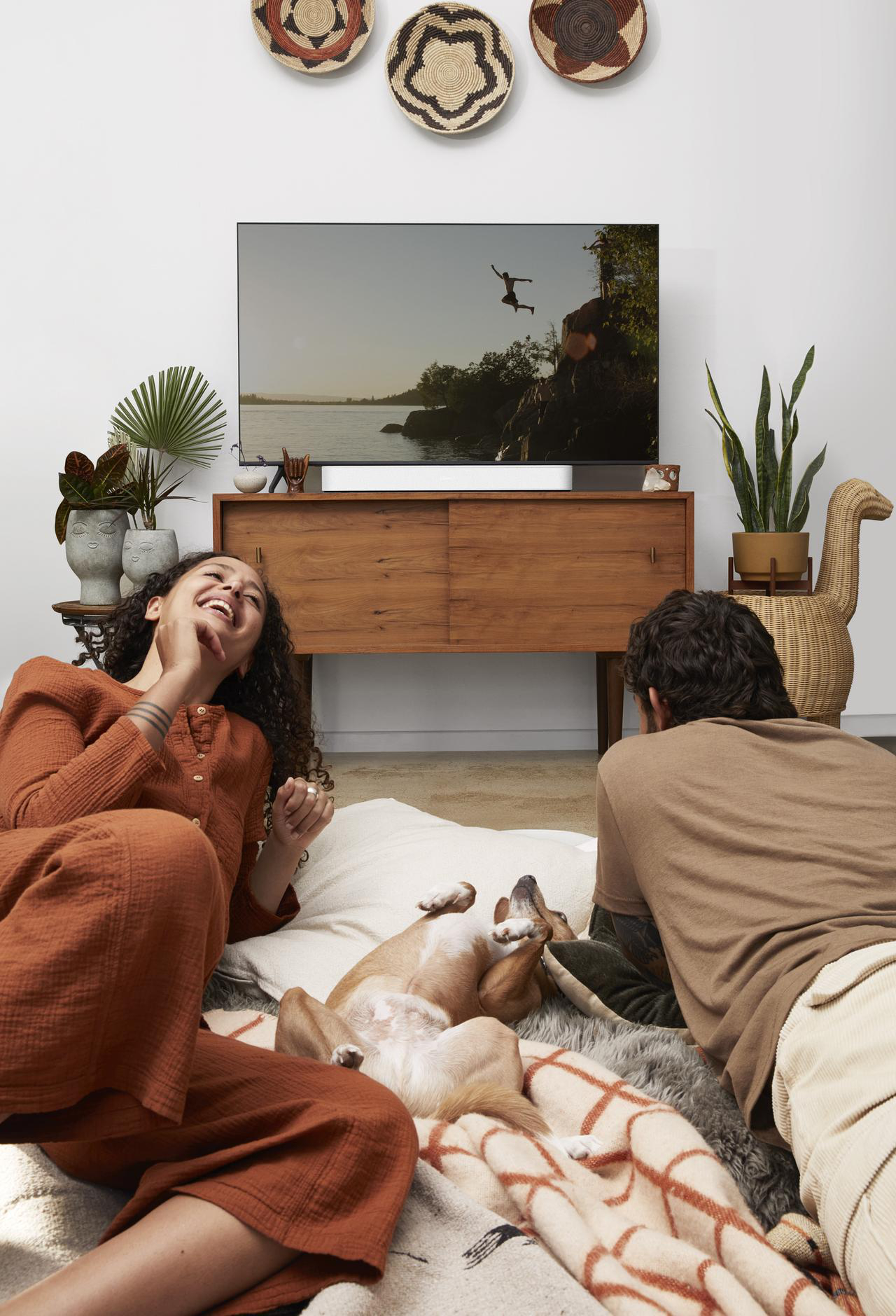 Menschen lachen, schauen fern, Hund dabei. Holzschrank und Pflanzen im Raum.