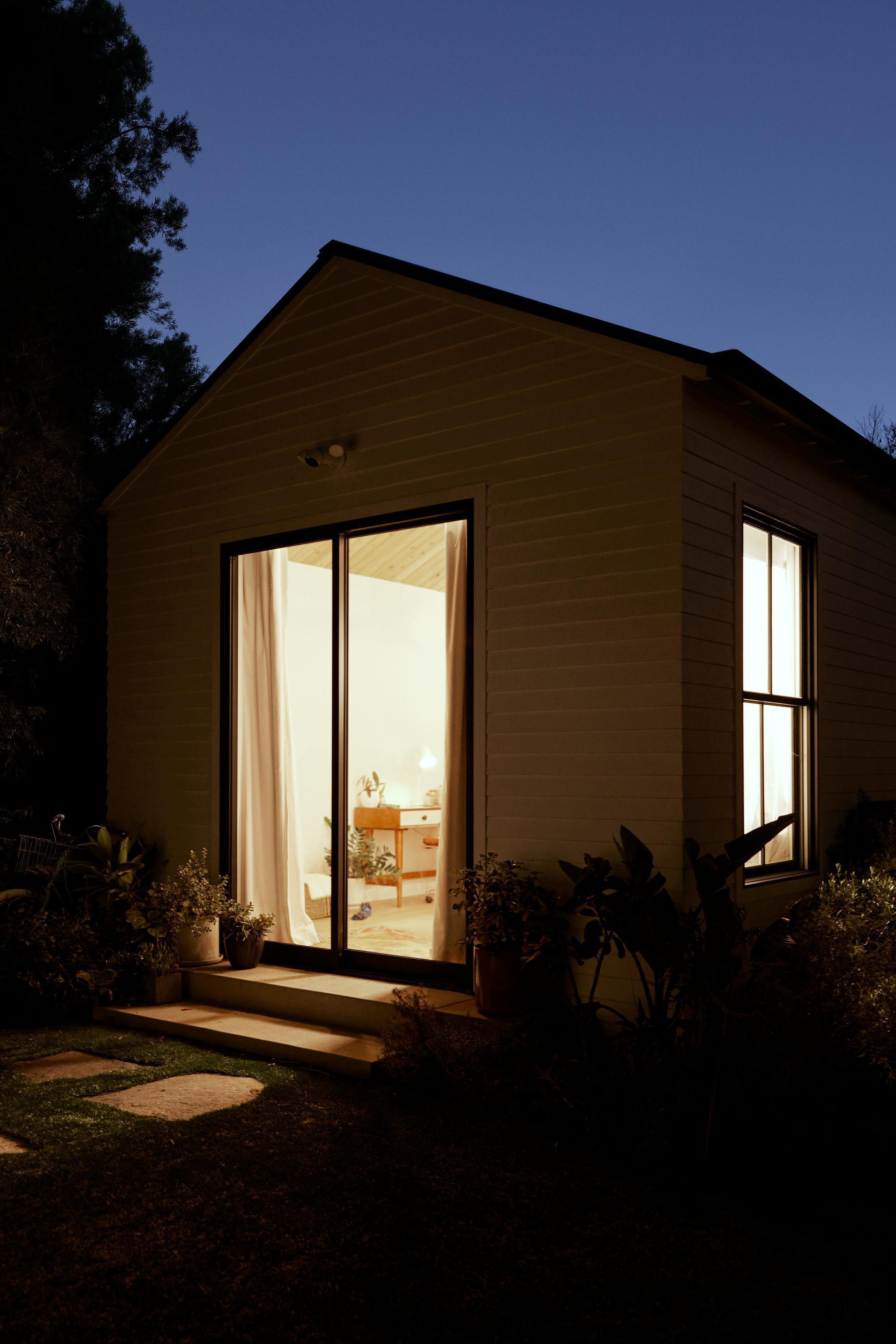 Ein kleines Haus in der Abenddämmerung mit Lichtern im Inneren, umgeben von Bäumen und Pflanzen.