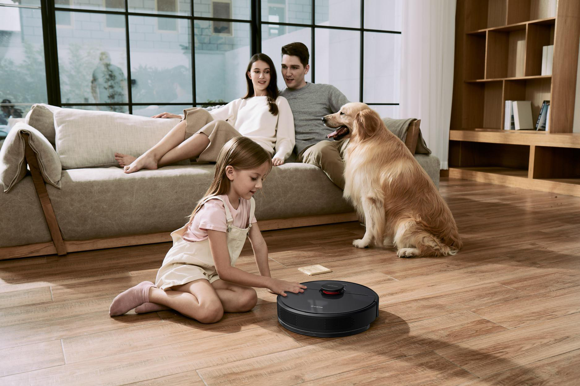 Eine Familie und ein Hund im Wohnzimmer, ein Mädchen spielt mit einem Roboterstaubsauger.