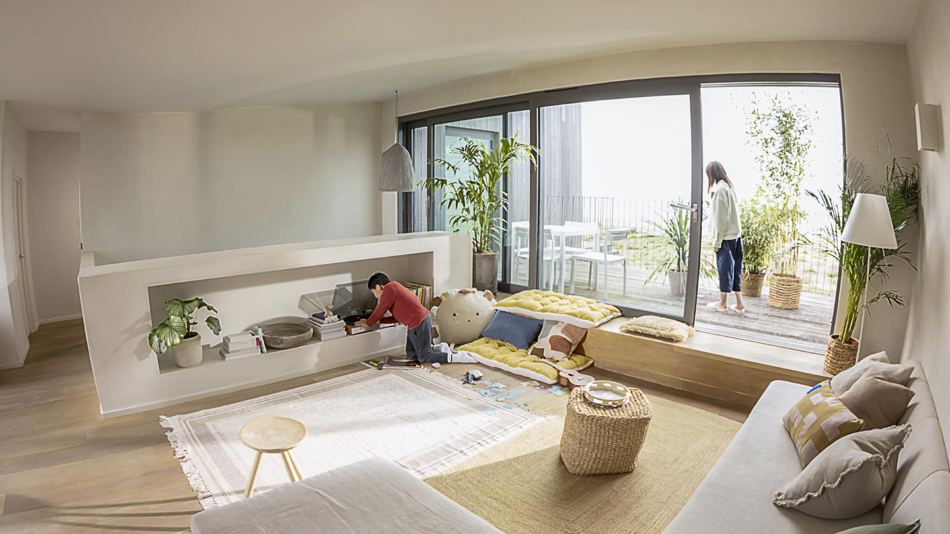 Ein Wohnzimmer mit spielendem Kind, Frau auf Balkon. Neutrale Töne und modernes Design.