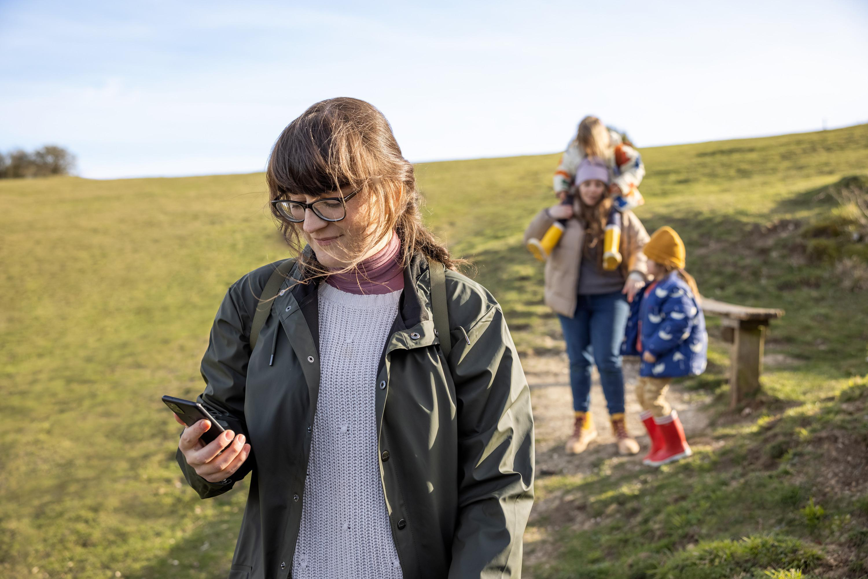 Eine Frau benutzt ihr Telefon auf einem grasbewachsenen Pfad mit einer Familie hinter sich.