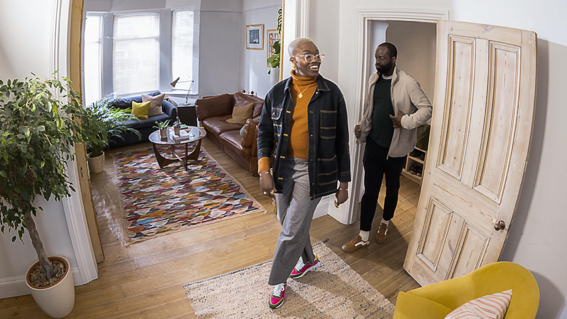 Zwei Personen in einem hellen Zuhause, eine geht auf die Kamera zu, die andere im Hintergrund. Moderne Einrichtung, Holzböden.