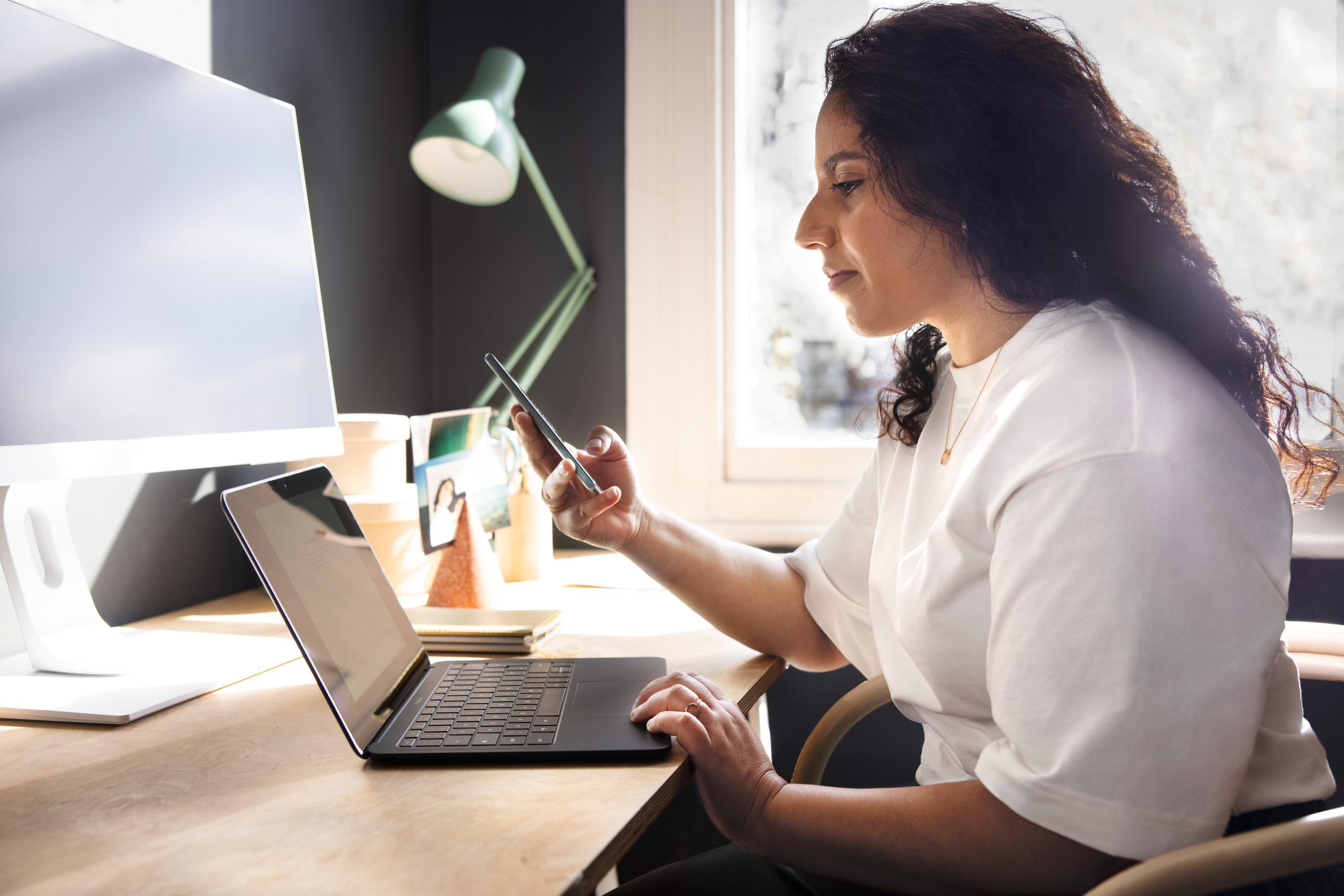 Frau, die ein Telefon benutzt, arbeitet am Laptop am Schreibtisch. Lampe und Monitor sichtbar.