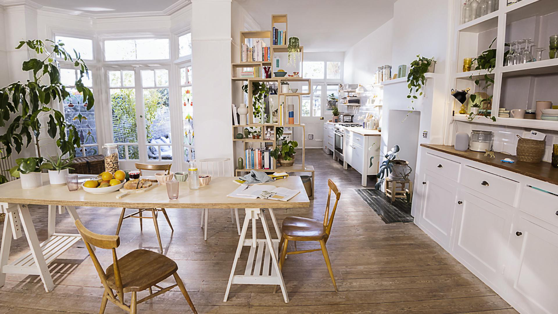Helle Küchenszene mit langem Tisch, Pflanzen und Bücherregal. Sonnenlicht durchflutet die Fenster.