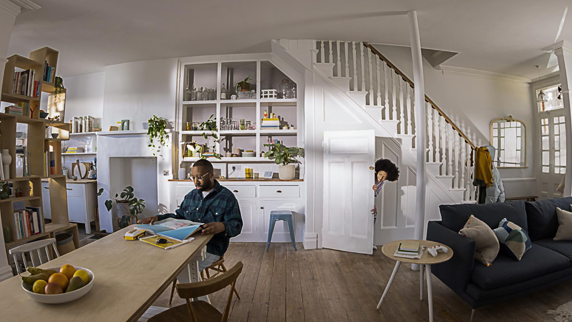 Ein Mann liest am Tisch, ein Kind lugt aus der Tür, ein Wohnzimmer.