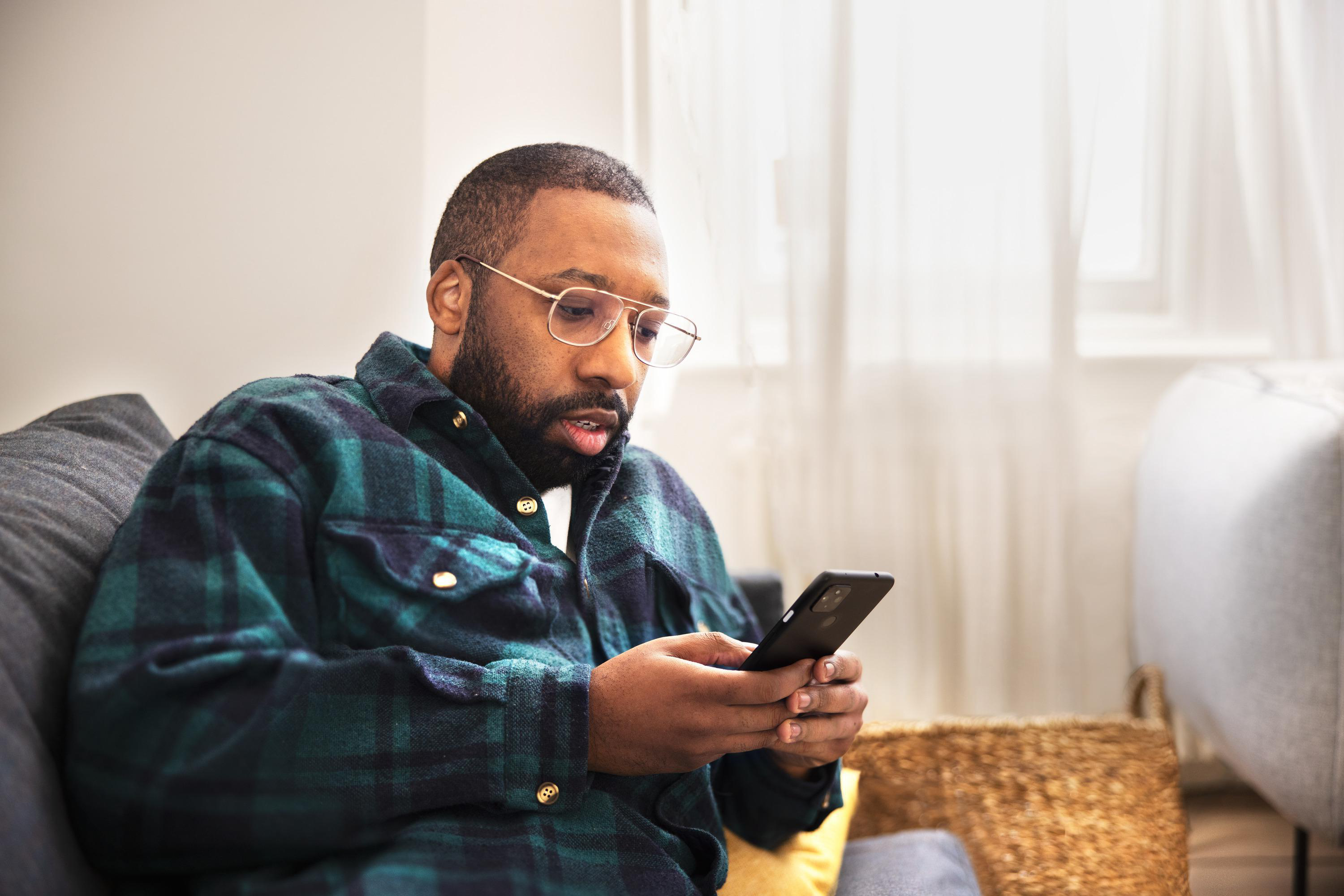Mann mit Brille schaut auf Handy auf Couch, grünes kariertes Hemd.