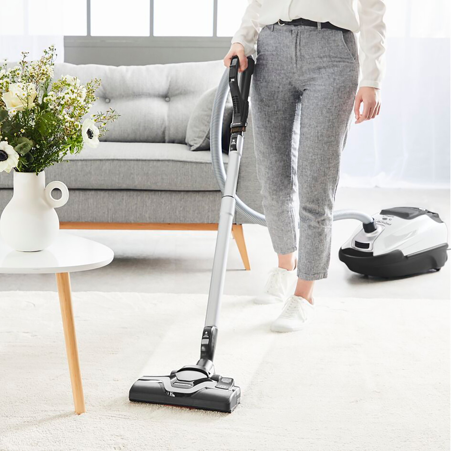 Vrouw die een wit tapijt stofzuigt in een woonkamer. Bank, tafel en vaas met bloemen zijn zichtbaar.