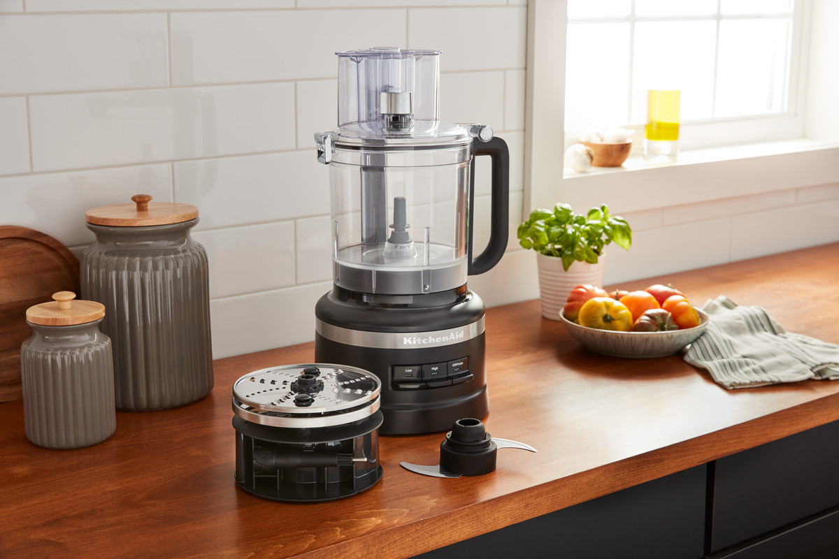 Ein KitchenAid-Küchenmaschine mit Teilen auf einer Theke, neben Gläsern und Basilikum.