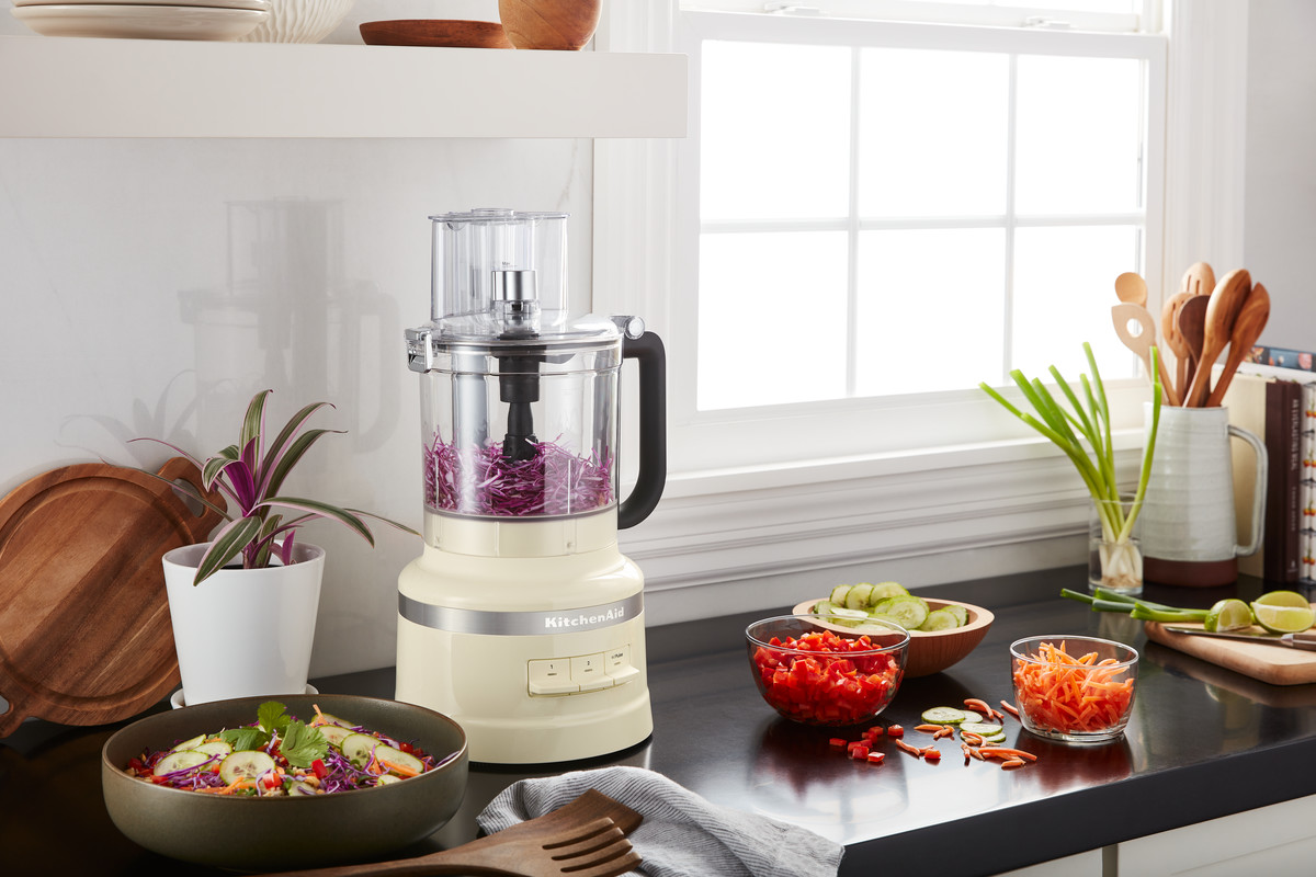 Ein cremefarbener KitchenAid-Foodprozessor mit Gemüse in Schalen und auf der Theke.