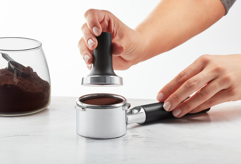 Hände, die Kaffeepulver in einen Siebträger stopfen. Ein Glas mit Pulver ist im Hintergrund.