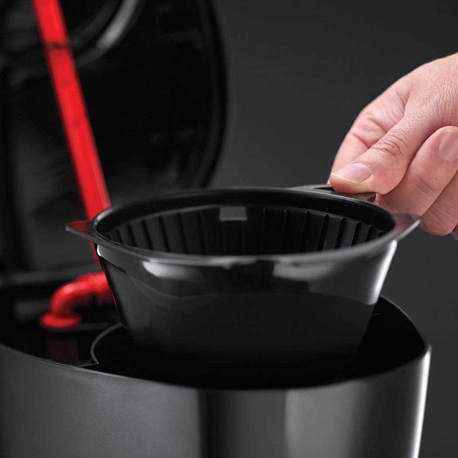 Una mano posiziona un cestello filtro per caffè nero in una macchina da caffè nera. Parti rosse sono visibili all'interno della macchina. Lo sfondo è scuro.