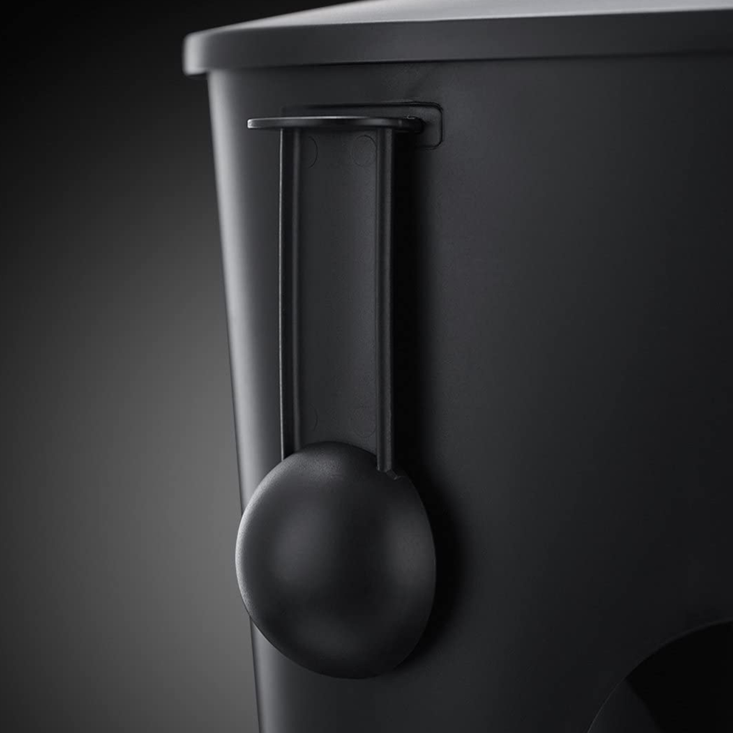 Una paletta di plastica nera è appesa a un contenitore nero. La paletta è rotonda. Lo sfondo è una sfumatura di grigio.