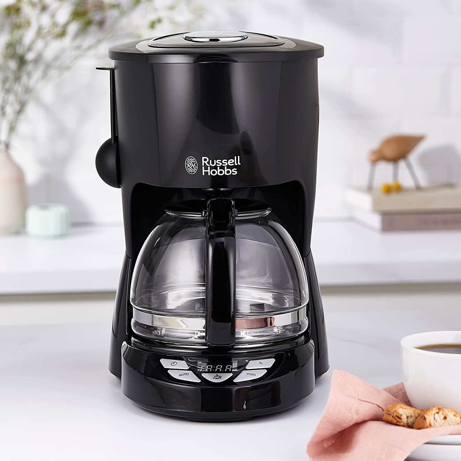 Caffettiera nera Russell Hobbs con caraffa in vetro, display digitale e tazza bianca con caffè. Tovagliolo rosa con biscotti.
