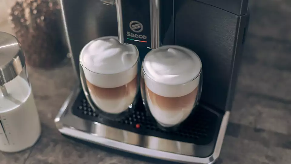 Two glasses of latte with foam, an espresso machine, and a milk container on a table.