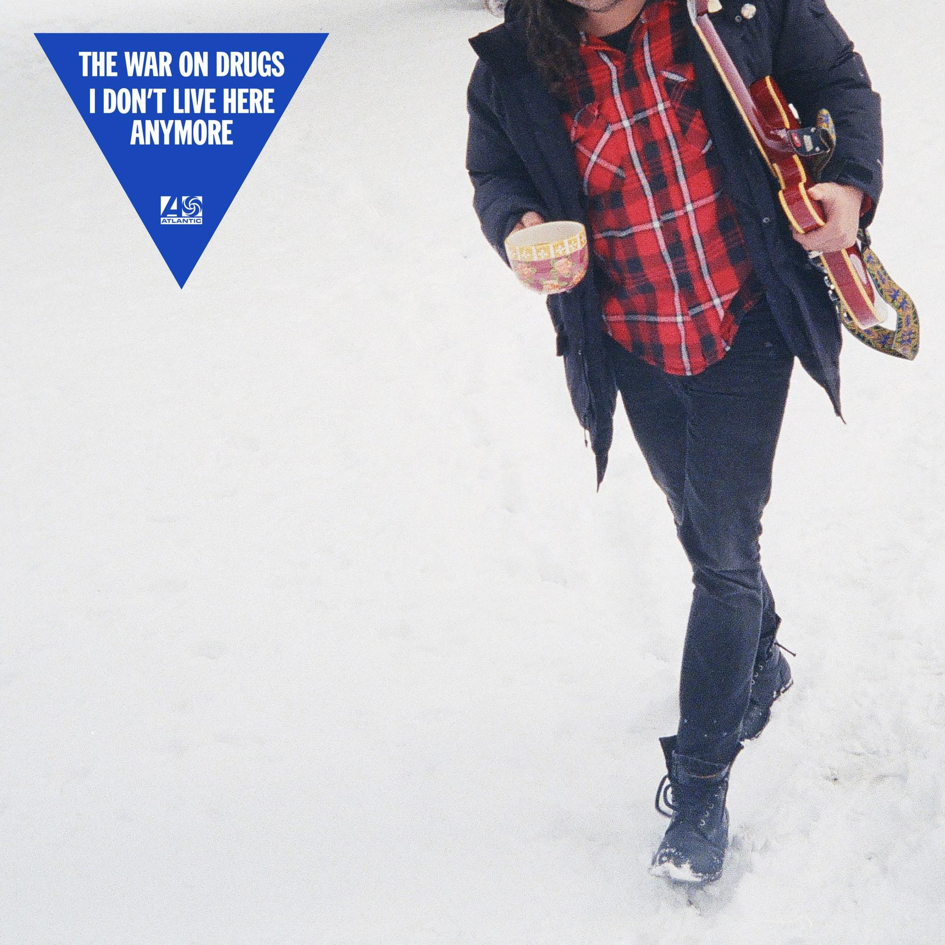 Homme marchant dans la neige. Il tient un bol et une guitare. Un panneau triangulaire bleu en haut à gauche.