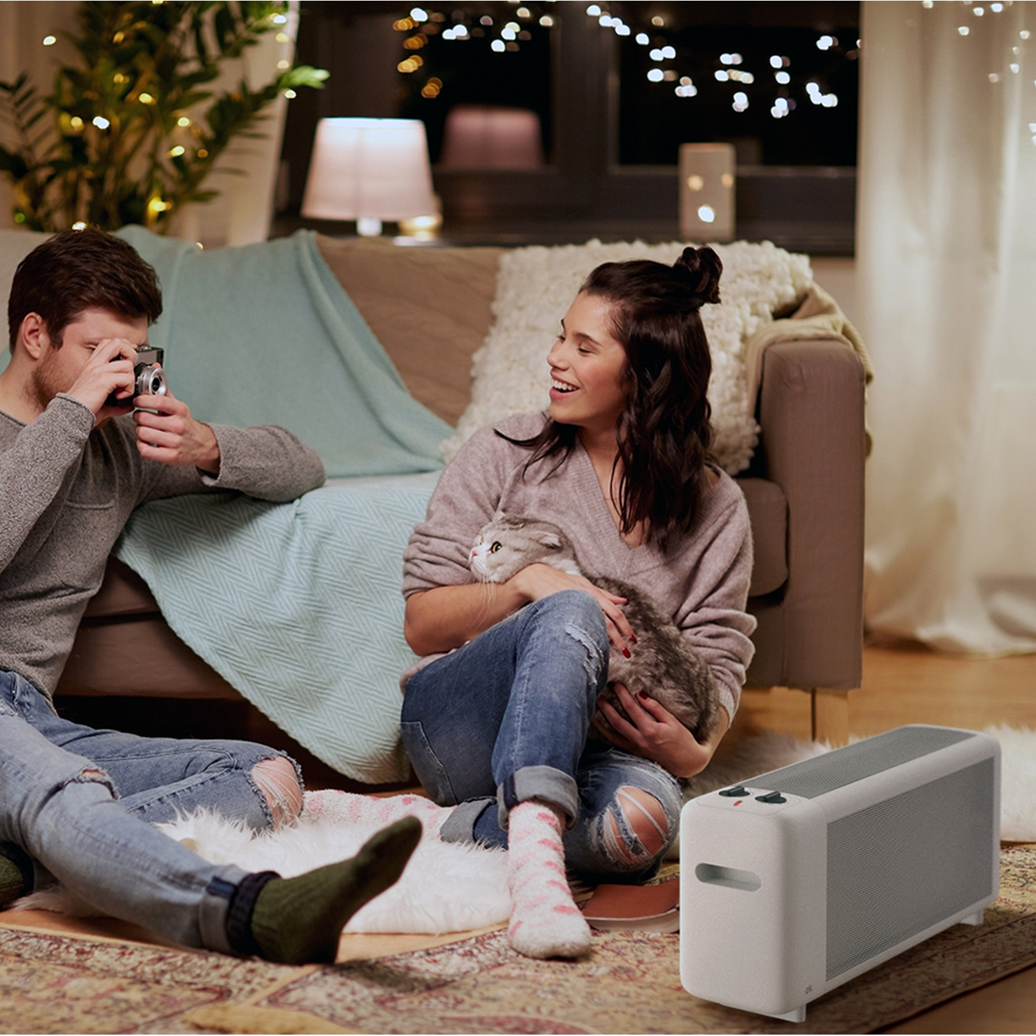 Una pareja y un gato en una sala de estar con un altavoz. Hombre tomando una foto. Mujer sosteniendo al gato.