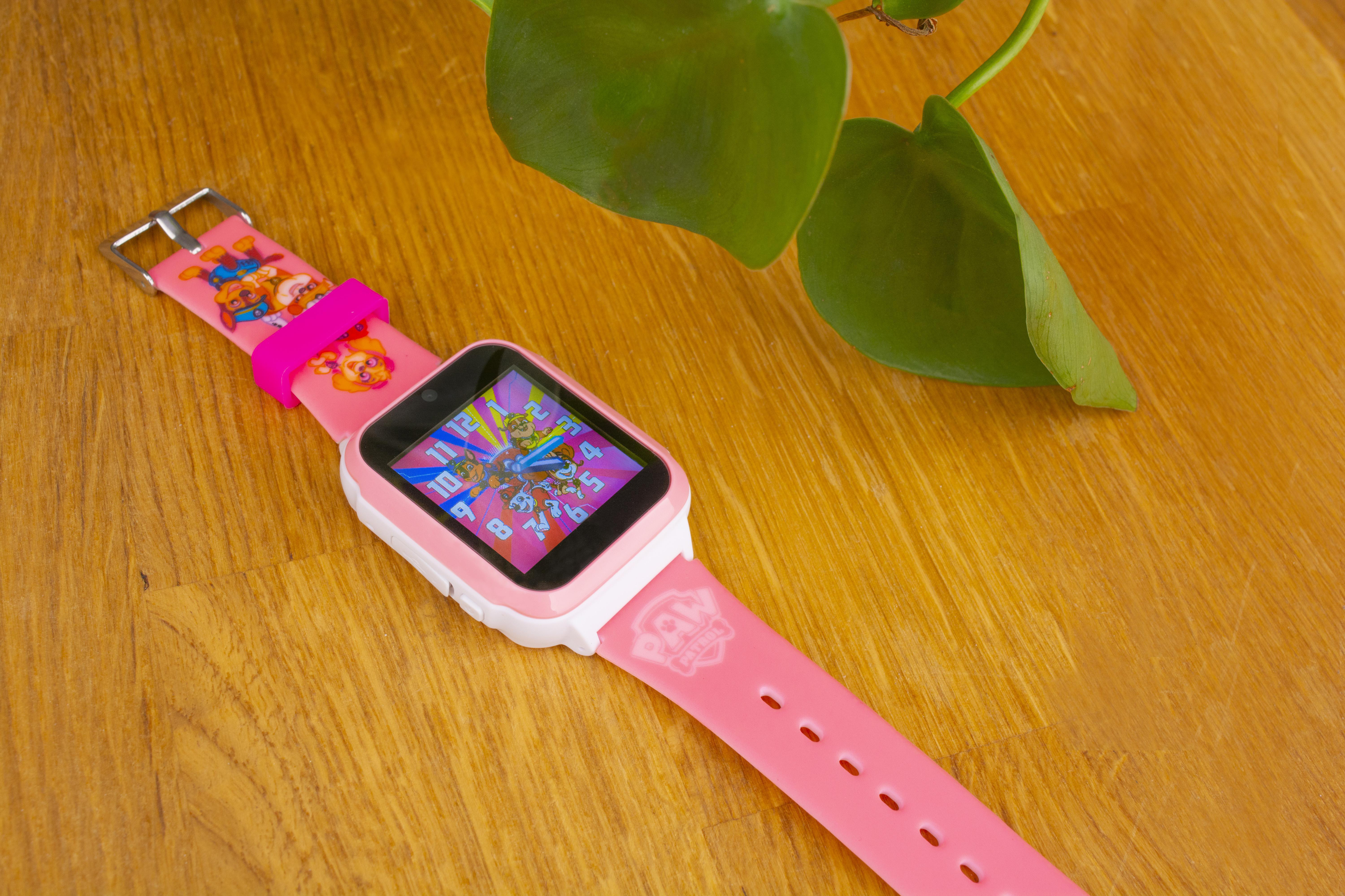 Eine pink-weiße Smartwatch mit Paw Patrol-Motiv, die auf einer Holzoberfläche mit einer grünen Pflanze liegt.