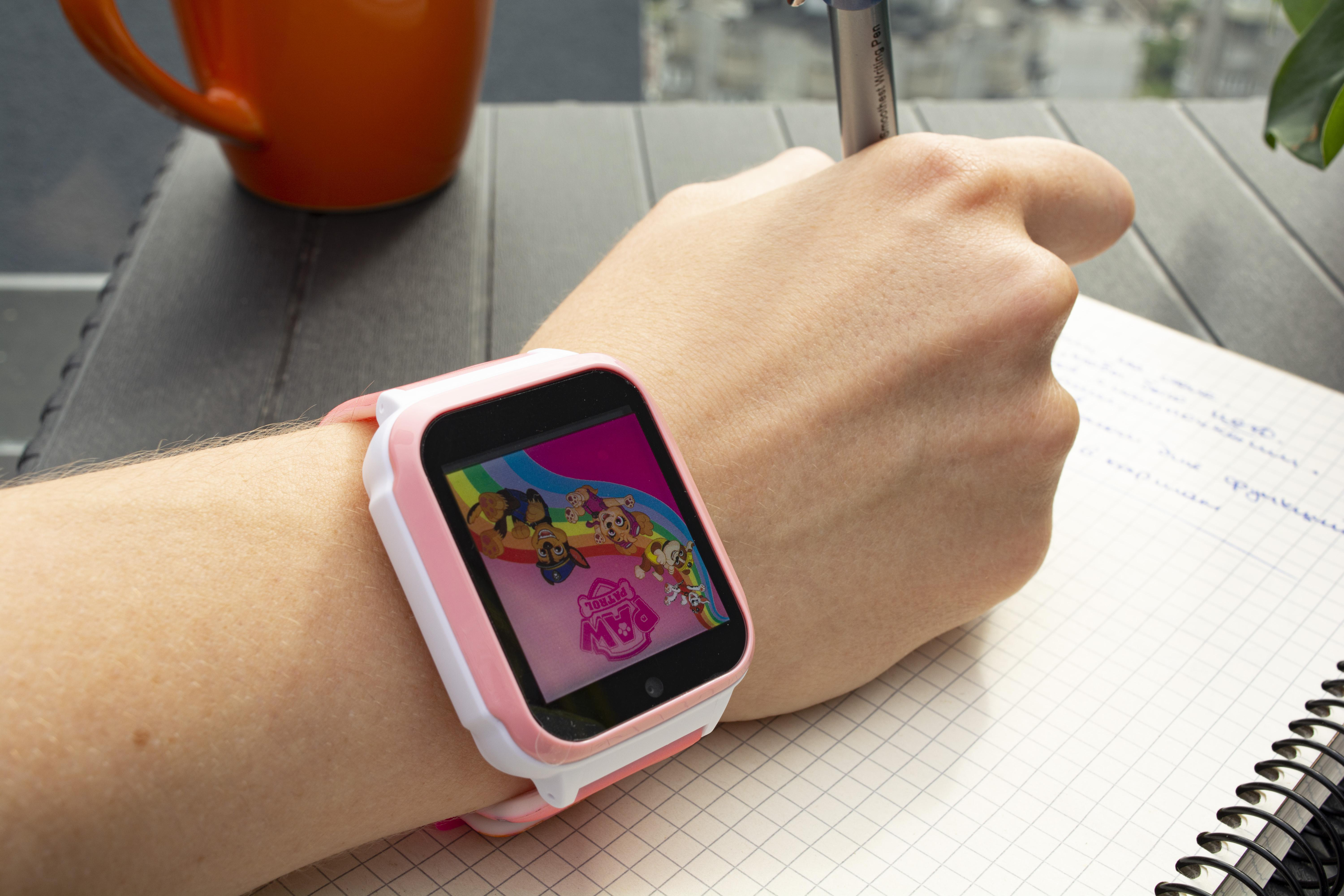 Handgelenk einer Person mit einer pink-weißen Smartwatch, die einen Cartoon zeigt. Stift und Papier daneben.