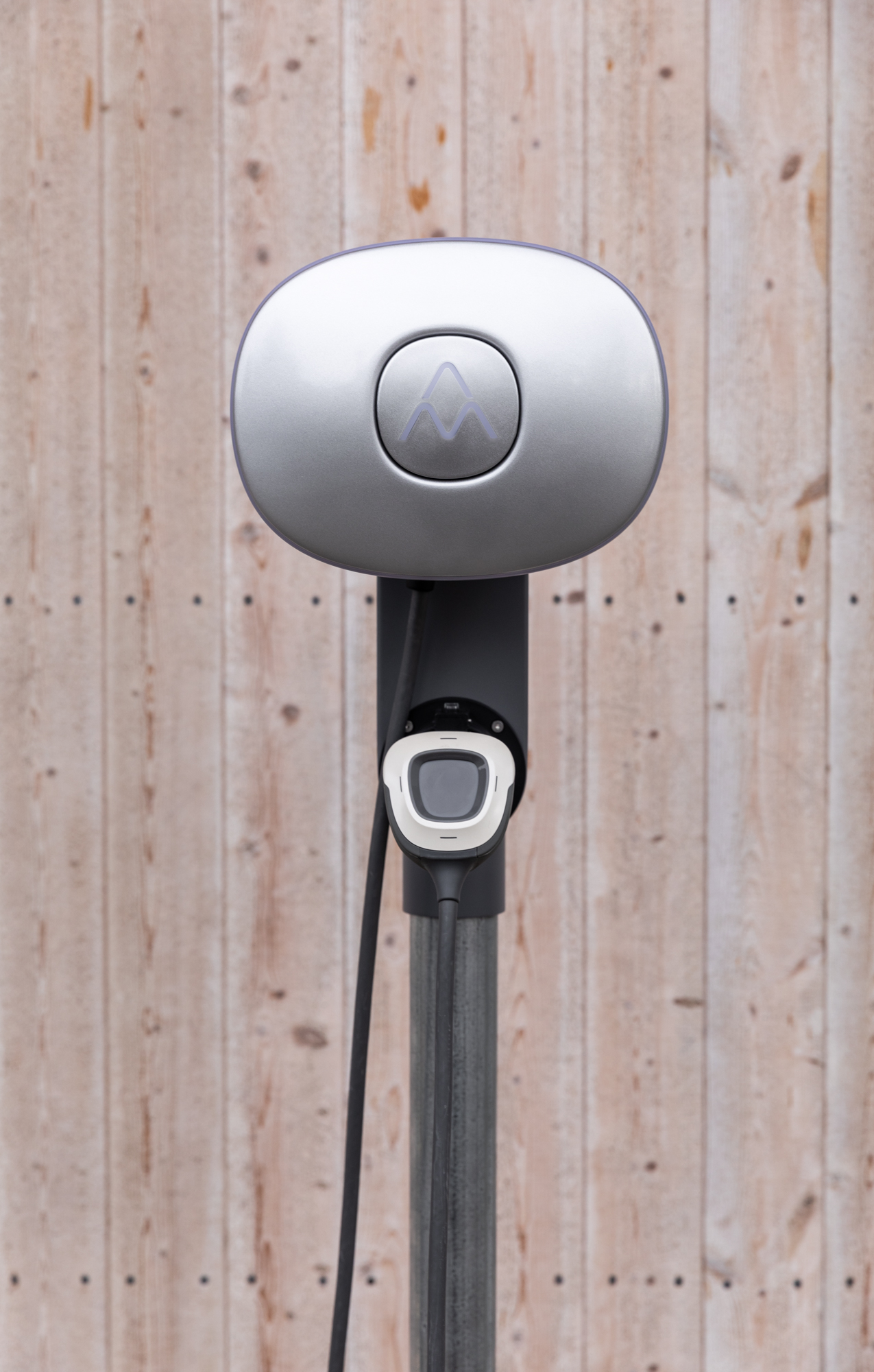 Eine silberne Ladestation für Elektrofahrzeuge mit einem schwarzen Kabel vor einer Holzwand.