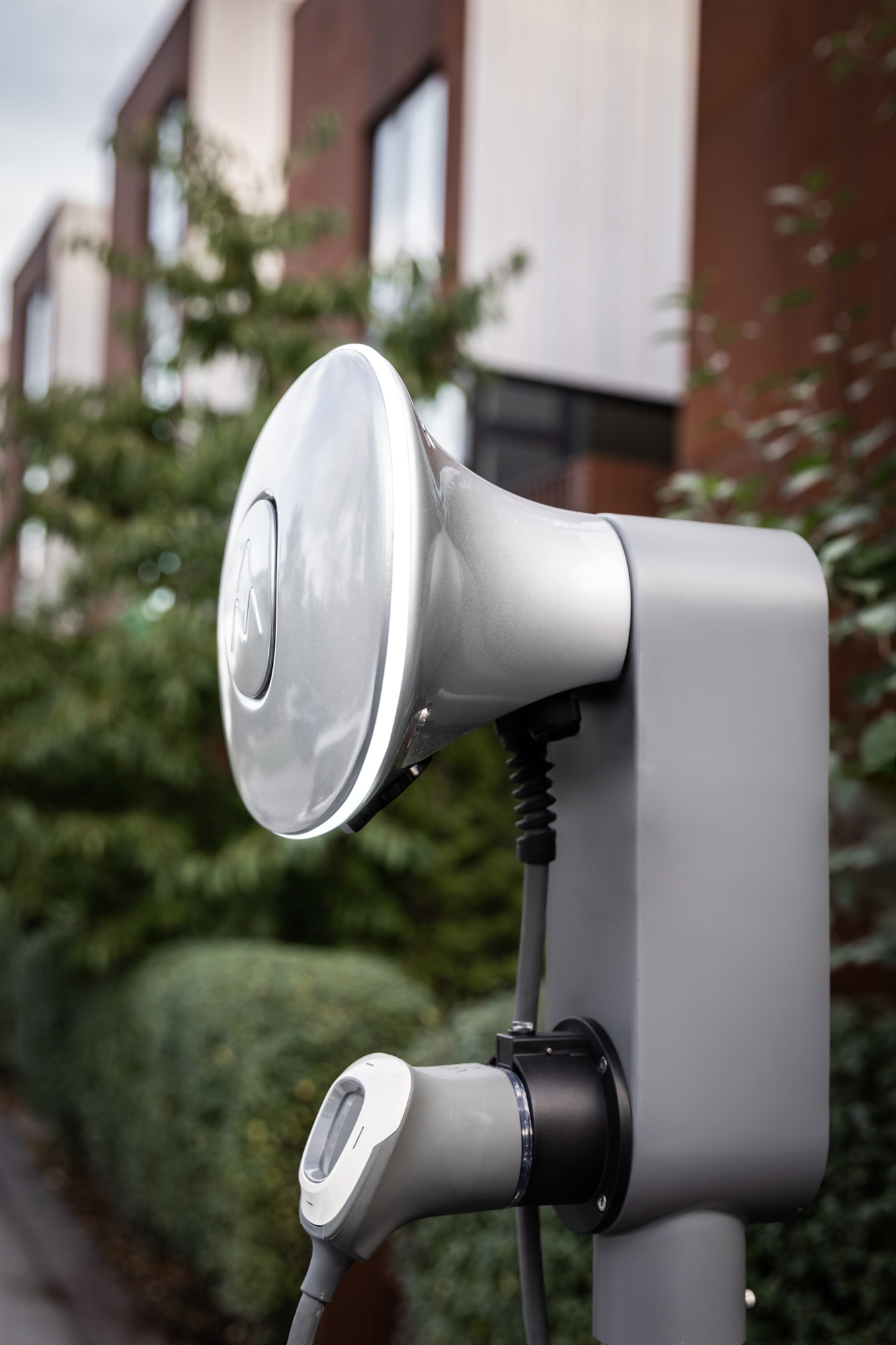 Eine silberne Ladestation für Elektrofahrzeuge mit Ladekabel und Gebäude im Hintergrund.