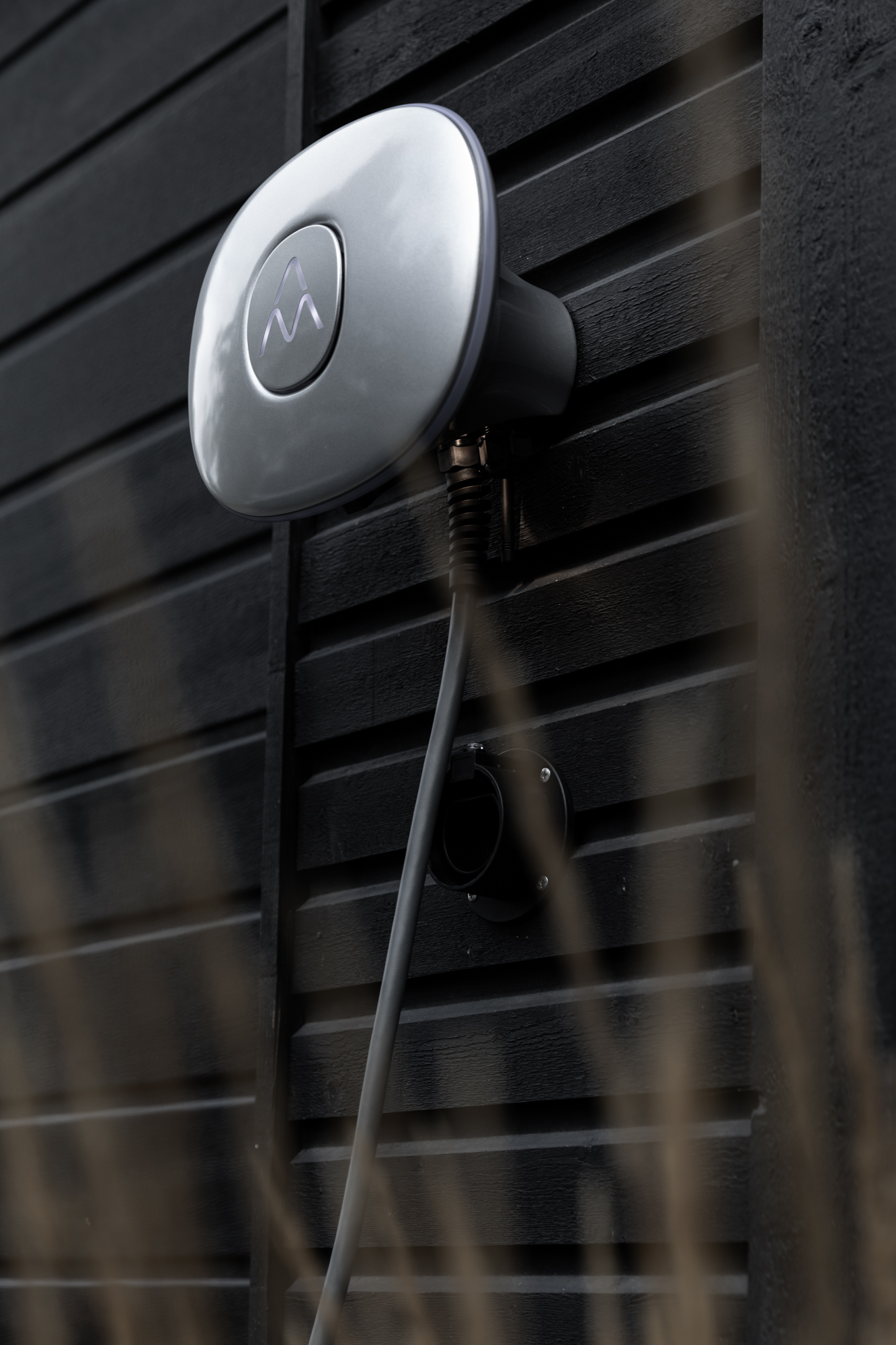 Eine EV-Ladestation an einer schwarzen Holzwand. Der Lader ist silberfarben und hat ein Logo.
