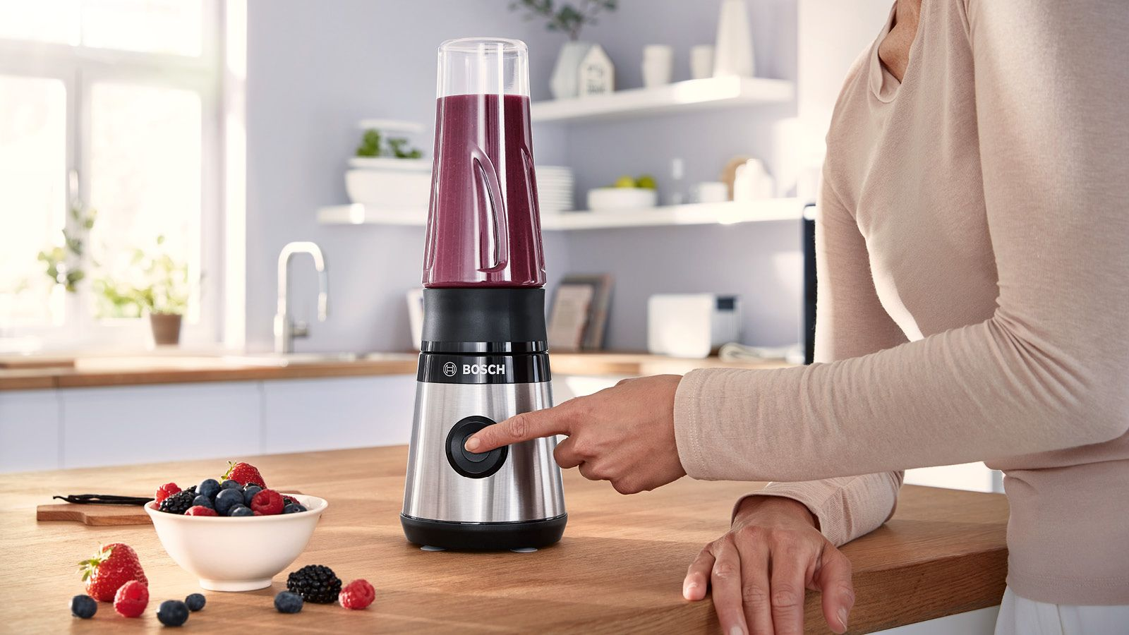 Egy nő megnyom egy gombot egy turmixgépen, egy pohár smoothie-val. Gyümölcsök a közelben.