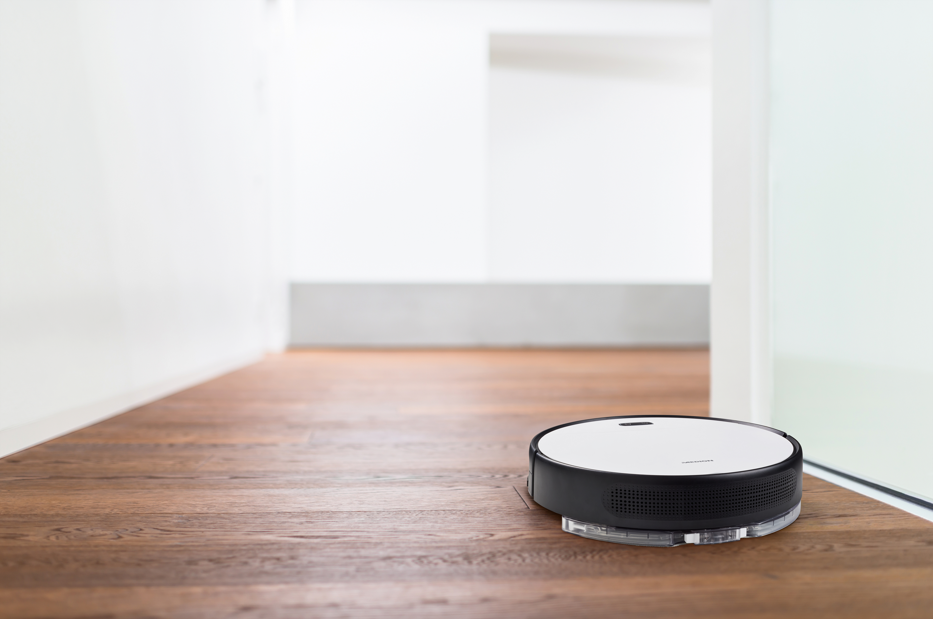 Ein Roboter-Staubsauger bewegt sich auf einem Holzboden in einem hellen, modernen Raum. Weiße Wände und Tür.