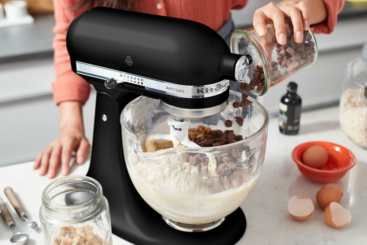 Ein schwarzer KitchenAid-Küchenmixer mit Zutaten. Eine Frau gibt Schokoladenstückchen hinzu.