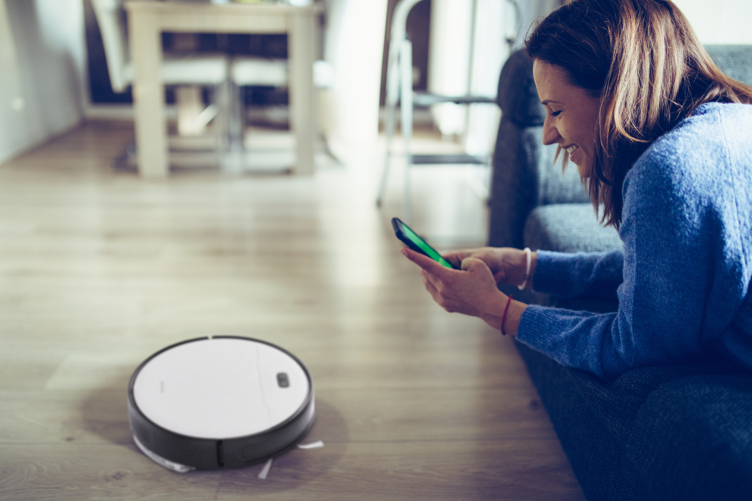 Eine Frau auf einem Sofa, die ein Telefon benutzt, während ein Roboter-Staubsauger den Boden reinigt.