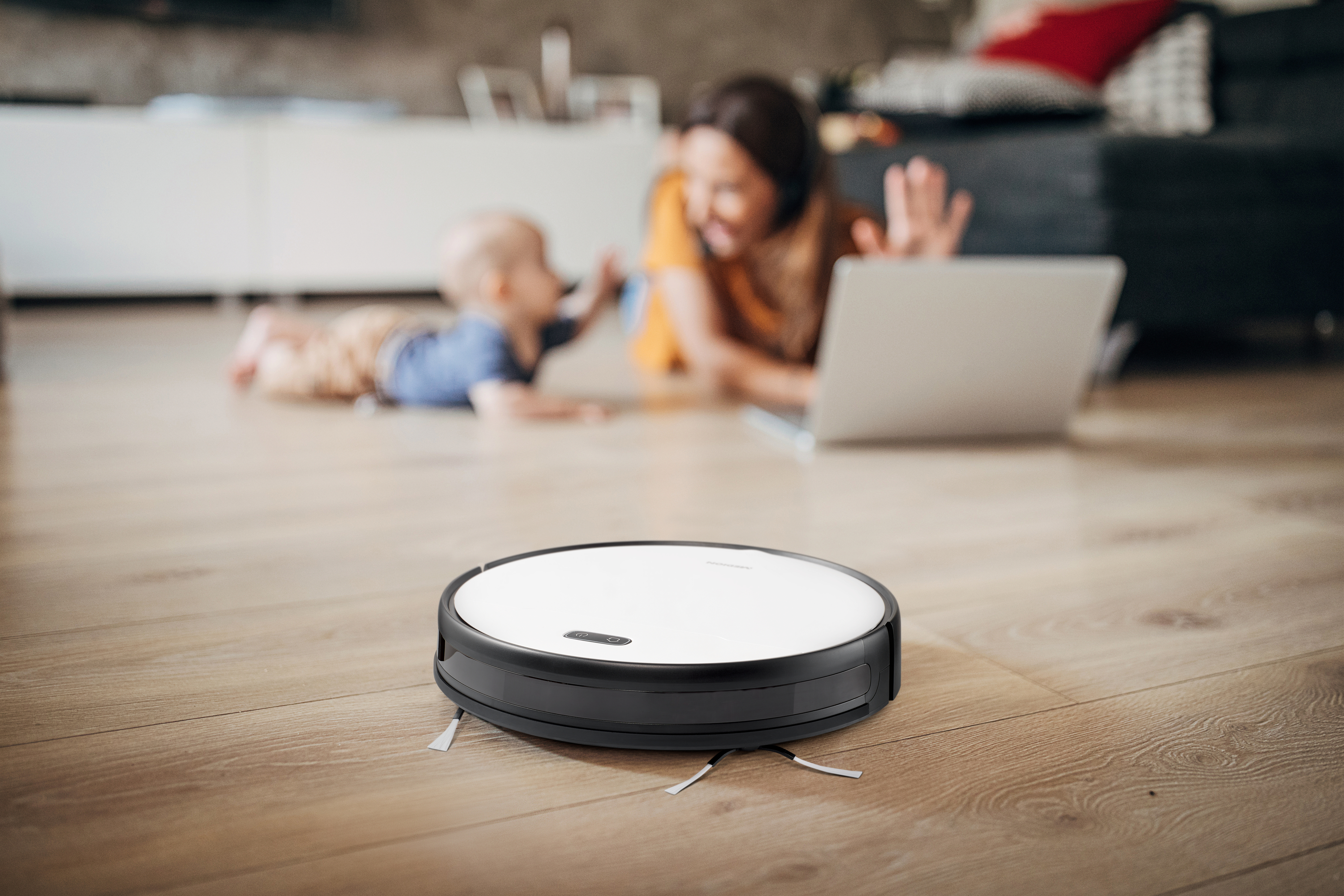 Roboter-Staubsauger auf Holzboden, Frau und Kind im Hintergrund.