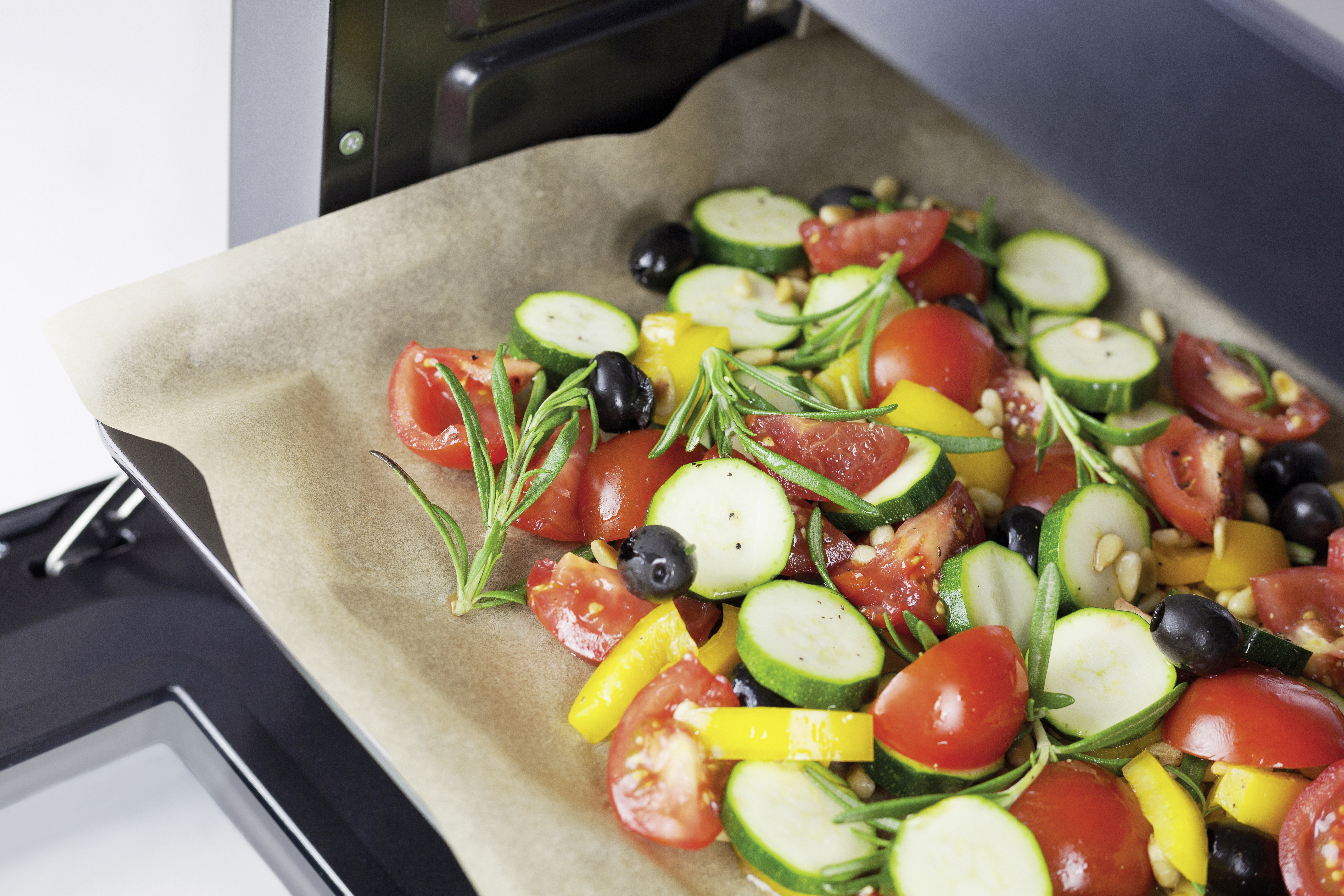 Ein Backblech mit geröstetem Gemüse: Tomaten, Zucchini, Oliven, Rosmarin und Pinienkerne.