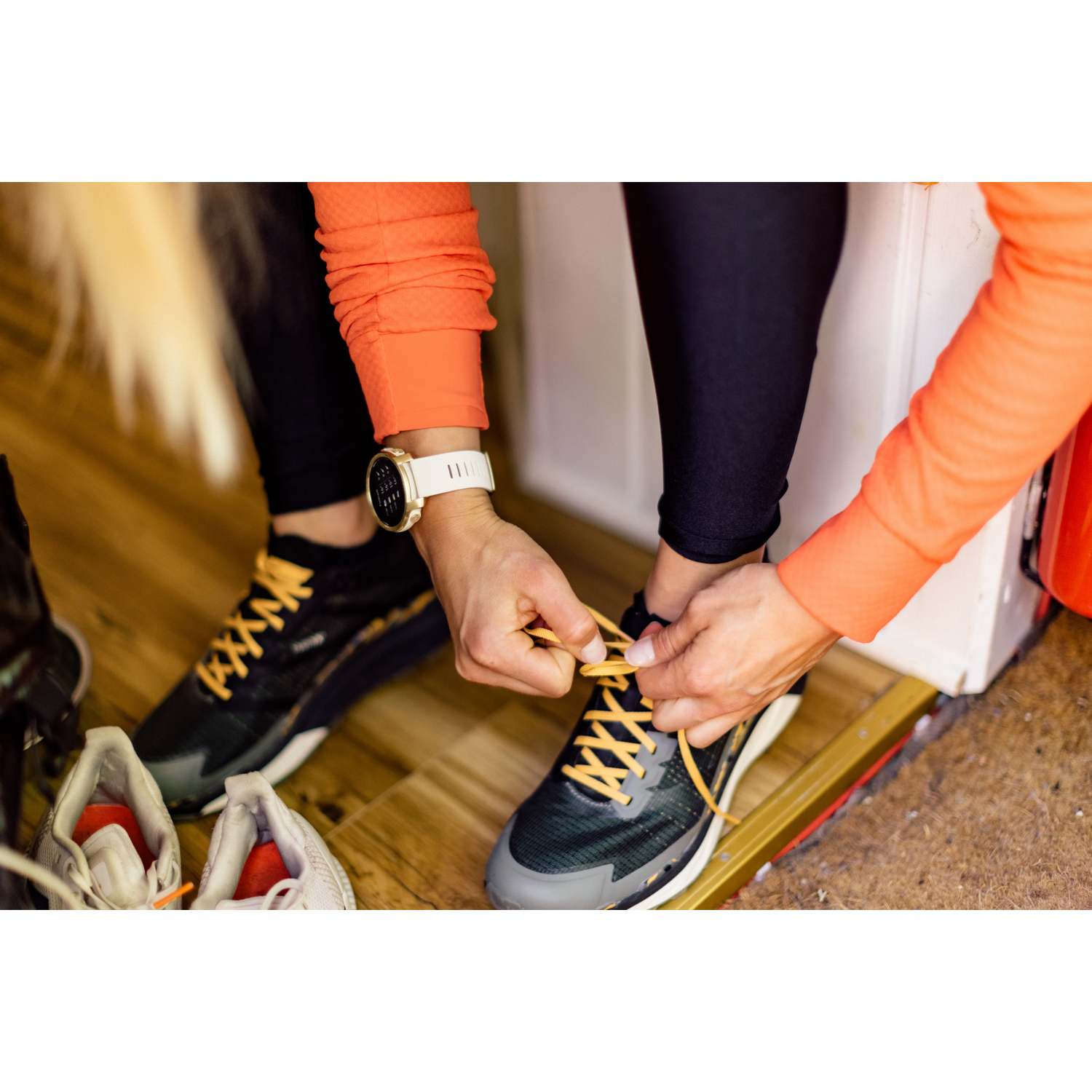 Una persona se ata los cordones. Llevan zapatillas para correr y un smartwatch. Otros zapatos están en el suelo.