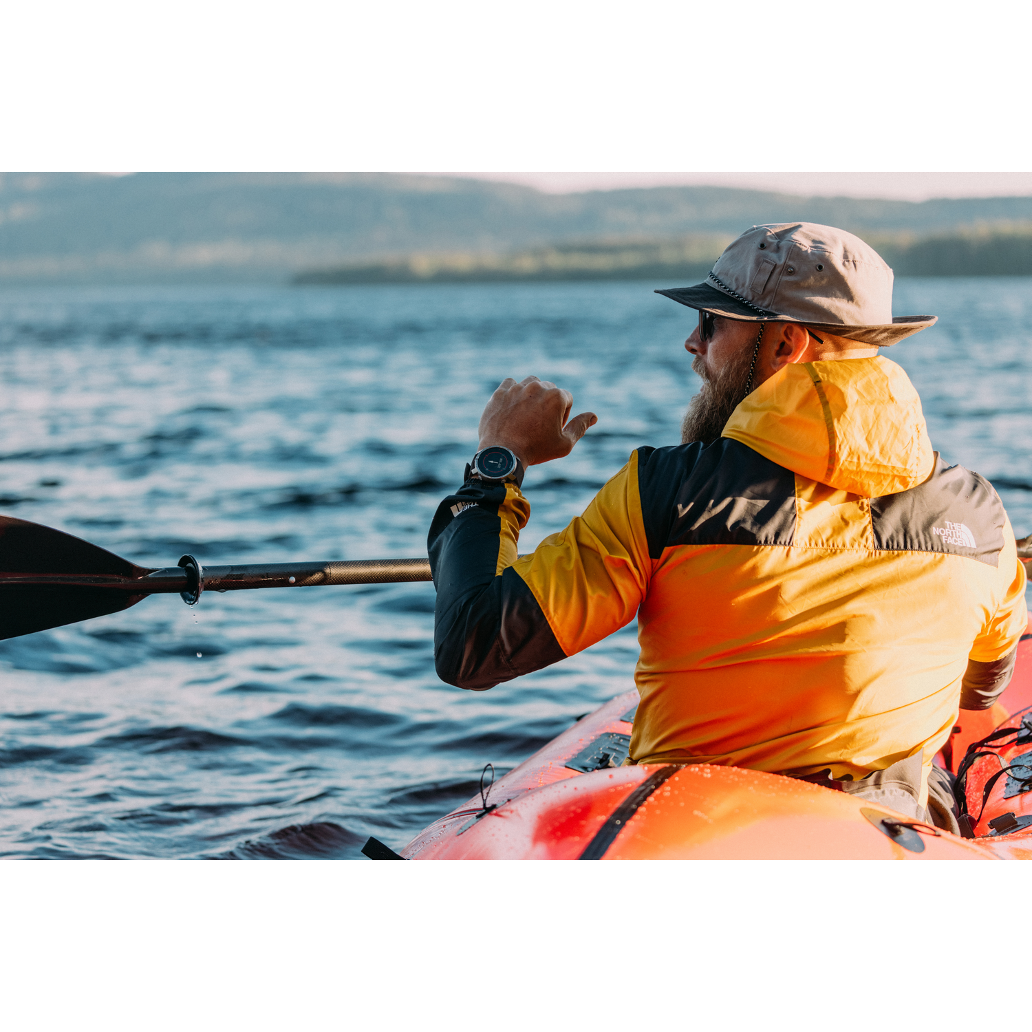 Un hombre en kayak en un lago, con sombrero y chaqueta, mirando su reloj.