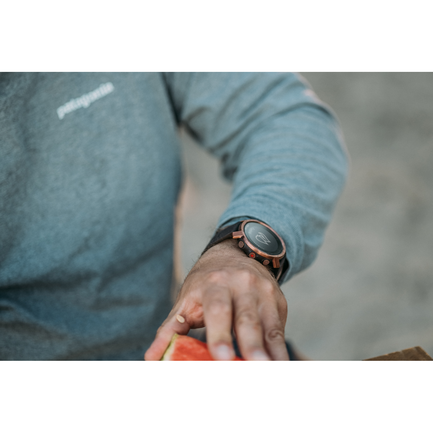 Persona con reloj, sosteniendo sandía, con fondo borroso al aire libre.