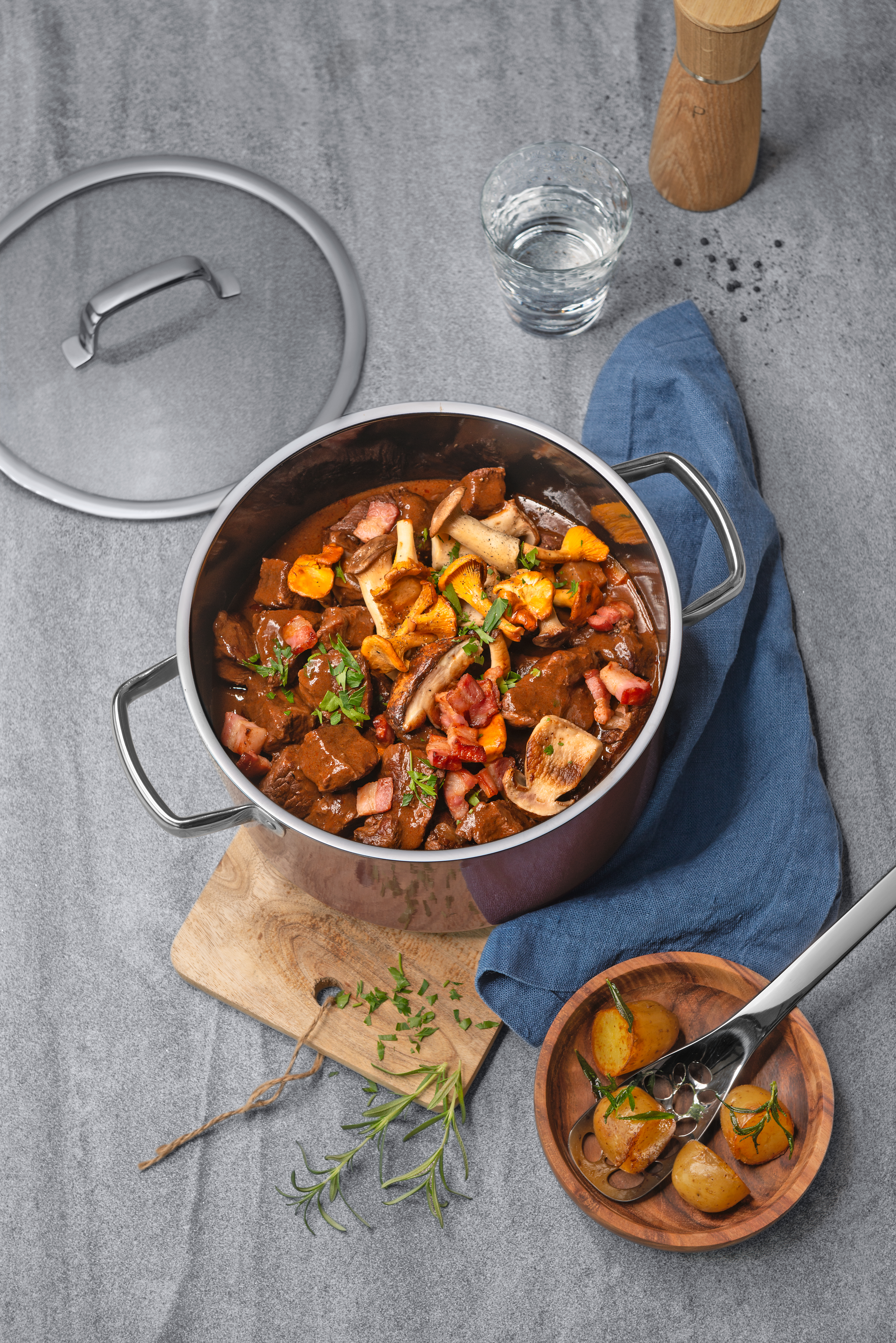 Eintopf in einem Kupfertopf mit Pilzen, Speck, Kartoffeln und Kräutern auf grauer Oberfläche.