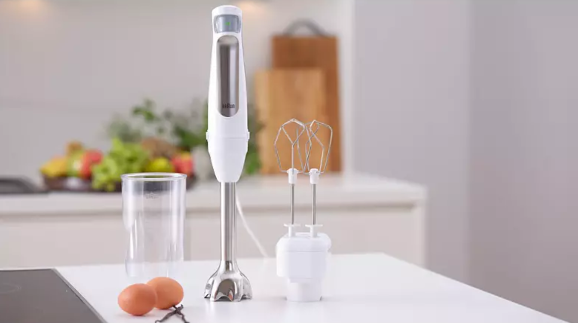 Mixeur plongeant blanc avec accessoires sur une table blanche. Le fond est une cuisine.