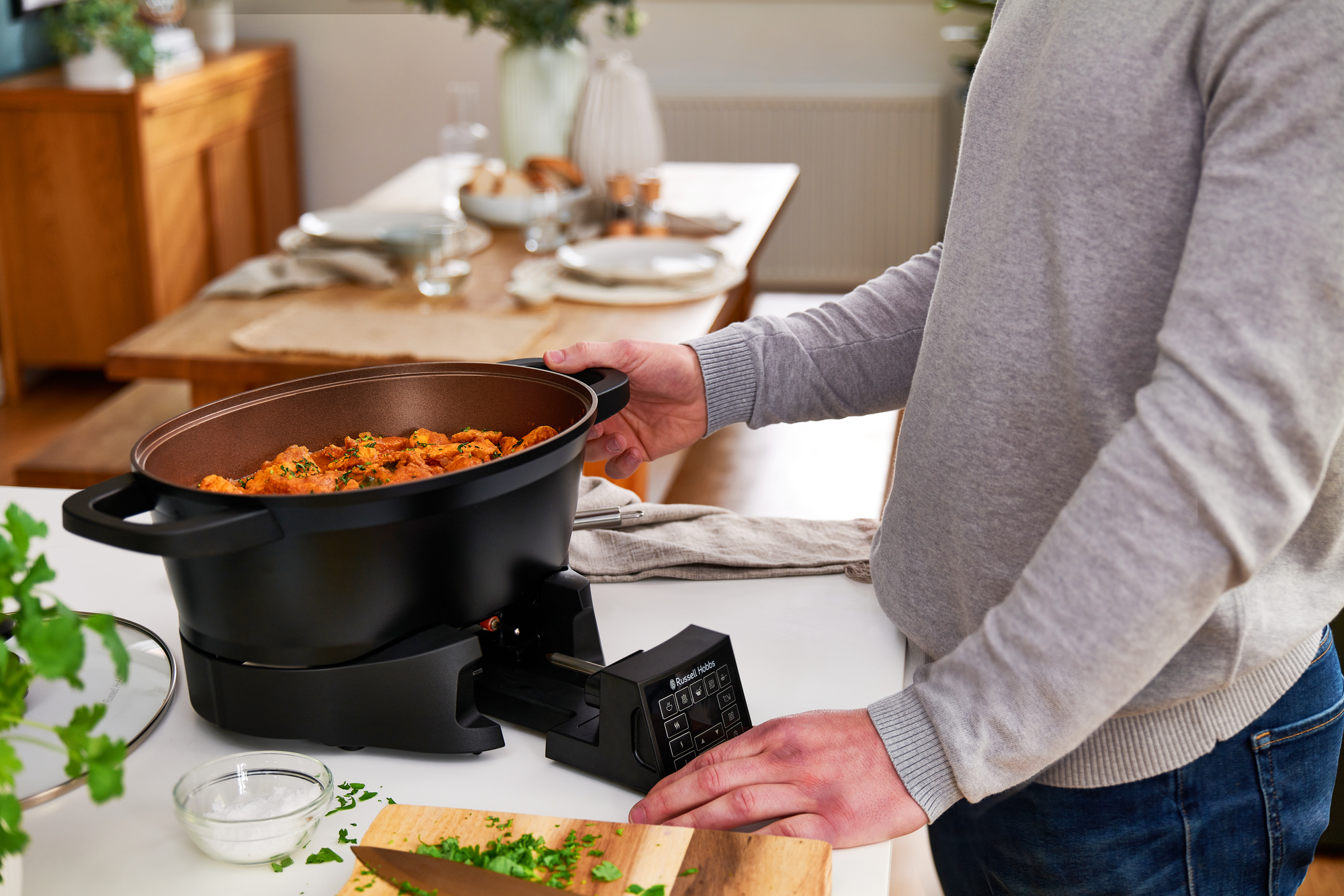 A person holding a black cooking pot.  The pot is on a black base next to some ingredients.
