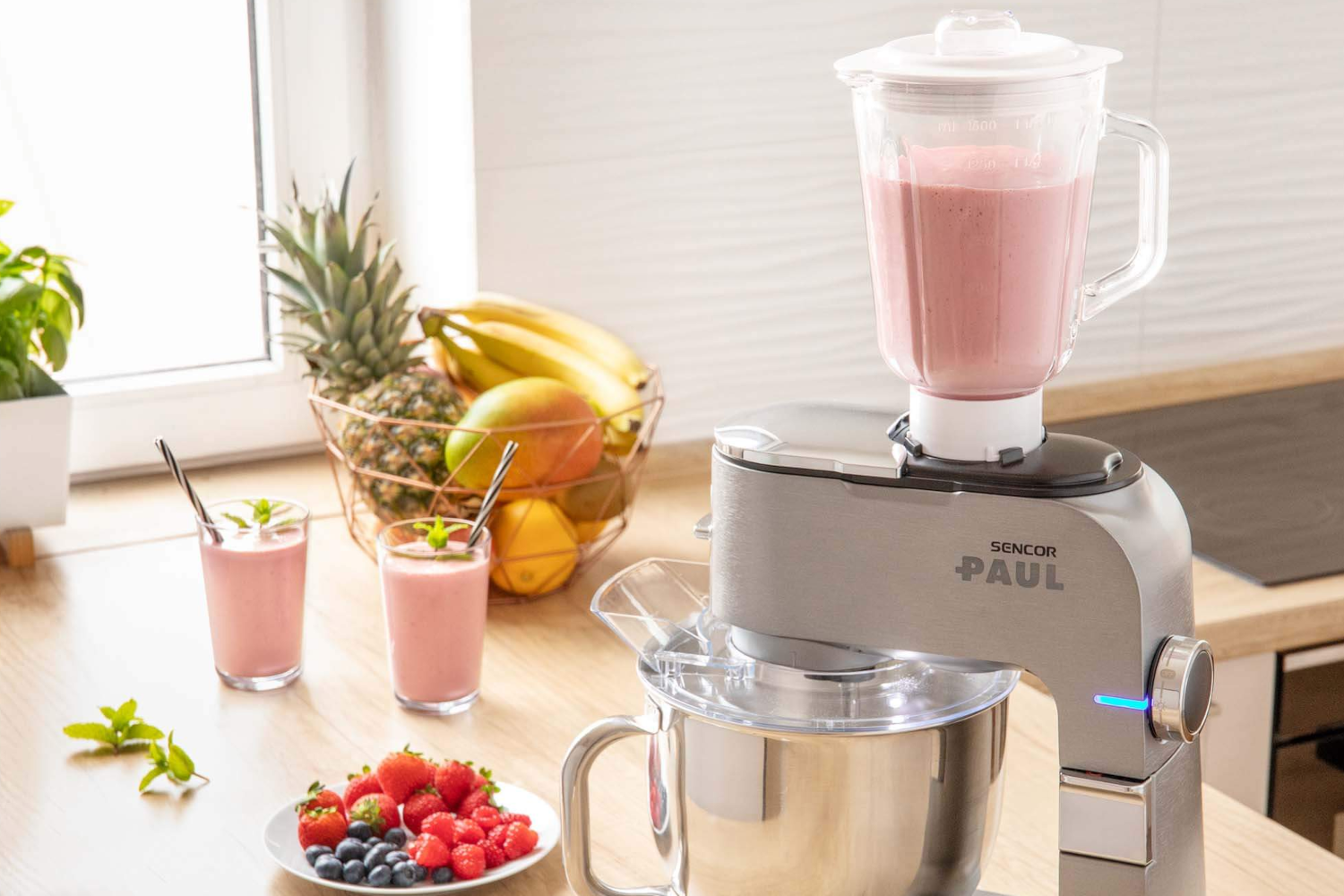 Egy turmixgép smoothie-val, gyümölcsökkel és két pohár rózsaszín smoothie-val.