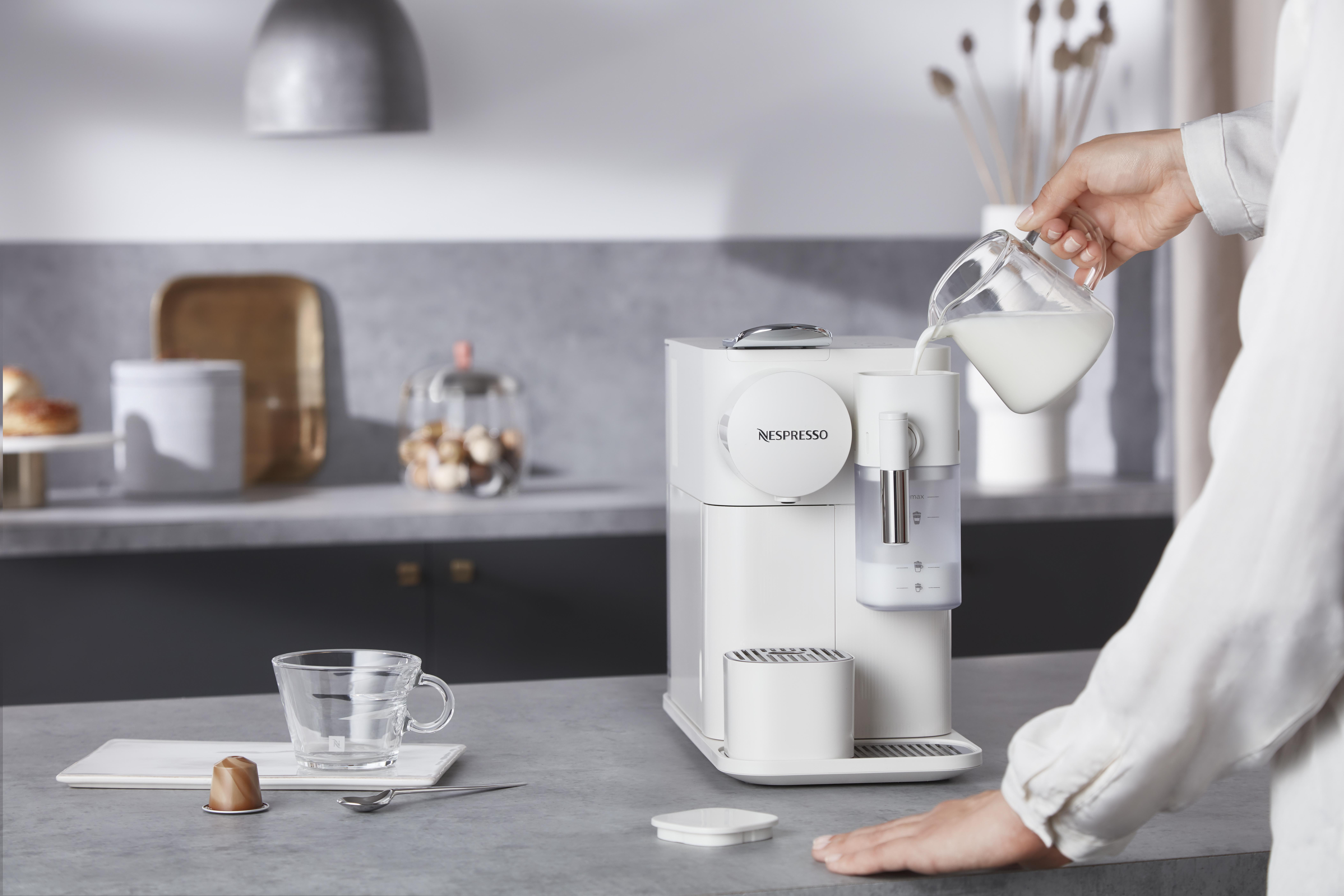 Eine Frau gießt Milch in eine Nespresso-Maschine. Weiße Maschine, Glas und Kapsel auf der Theke.