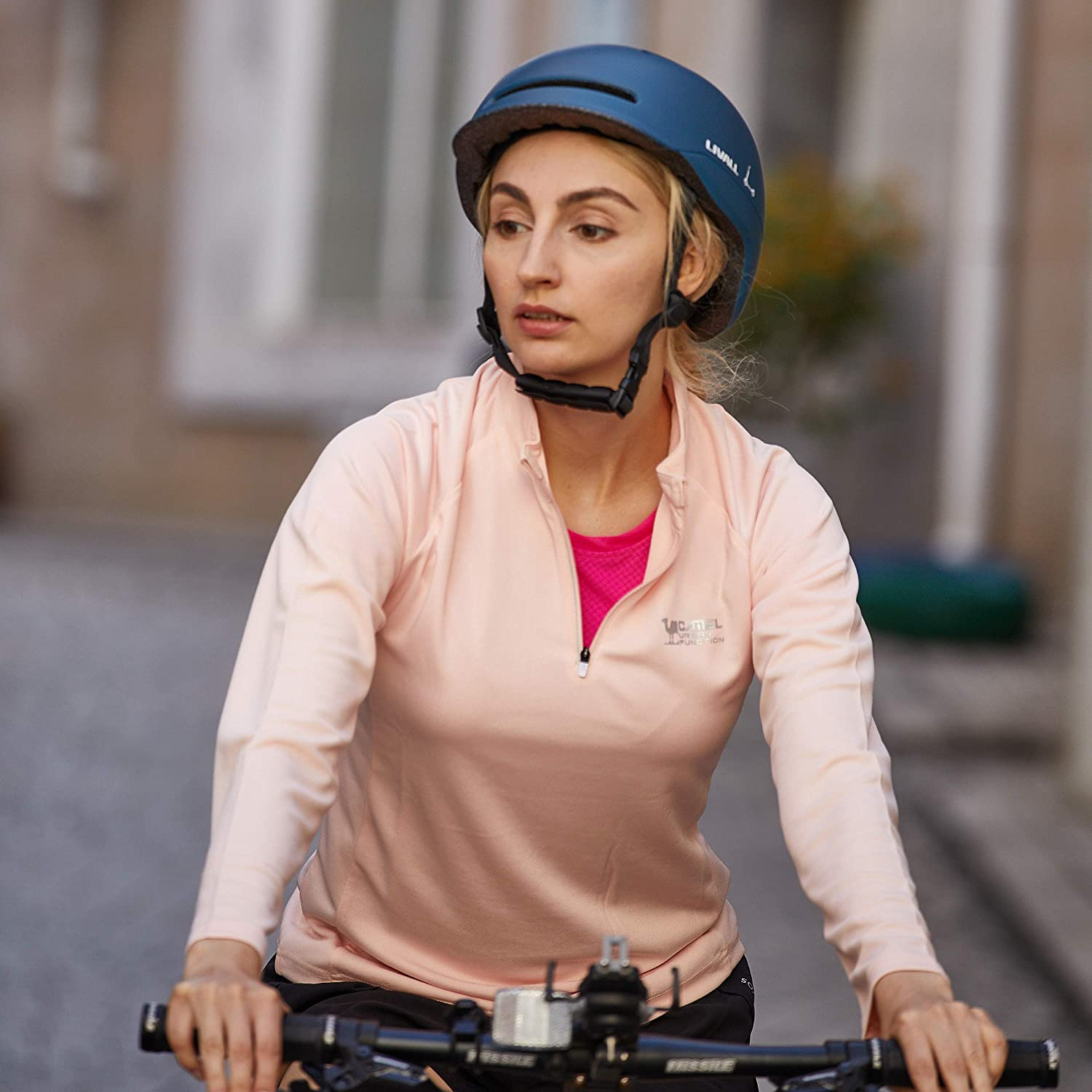 Mujer montando en bicicleta, con casco y top rosa, circulando por una calle de la ciudad.