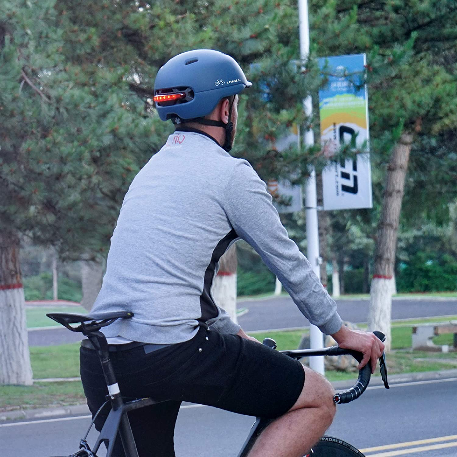 Ciclista con casco azul con luces traseras, montando en bicicleta en una carretera con árboles.