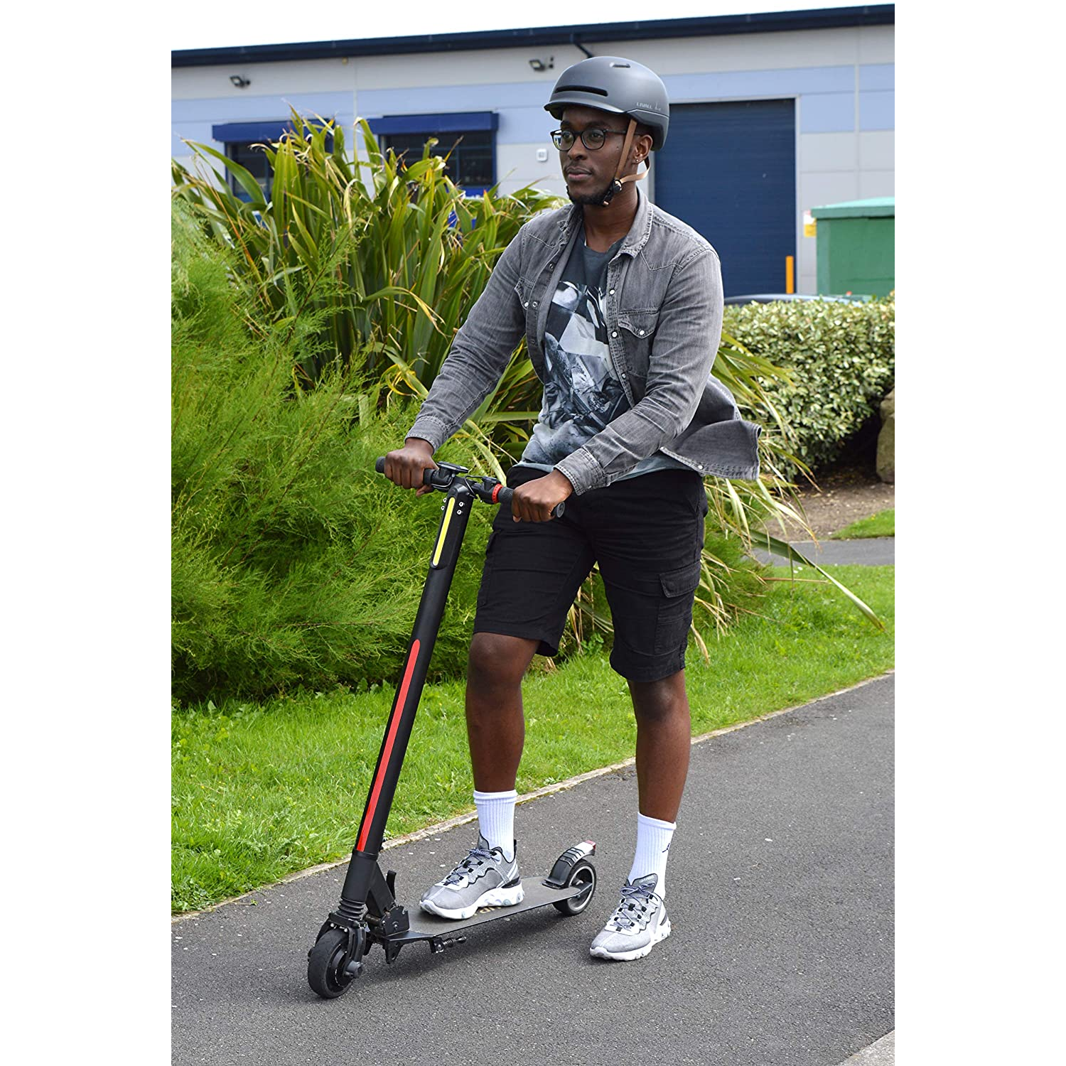 Un hombre con casco y gafas montando un patinete eléctrico en un camino. Edificios en el fondo.