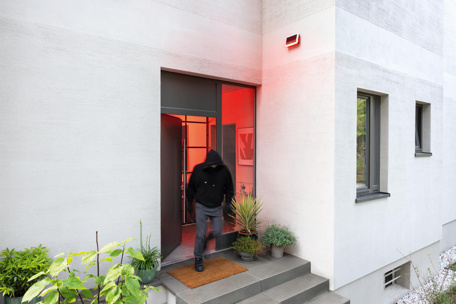 Eine Person in einem Kapuzenpullover rennt aus einem weißen Gebäude mit roter Beleuchtung. Tür ist offen.