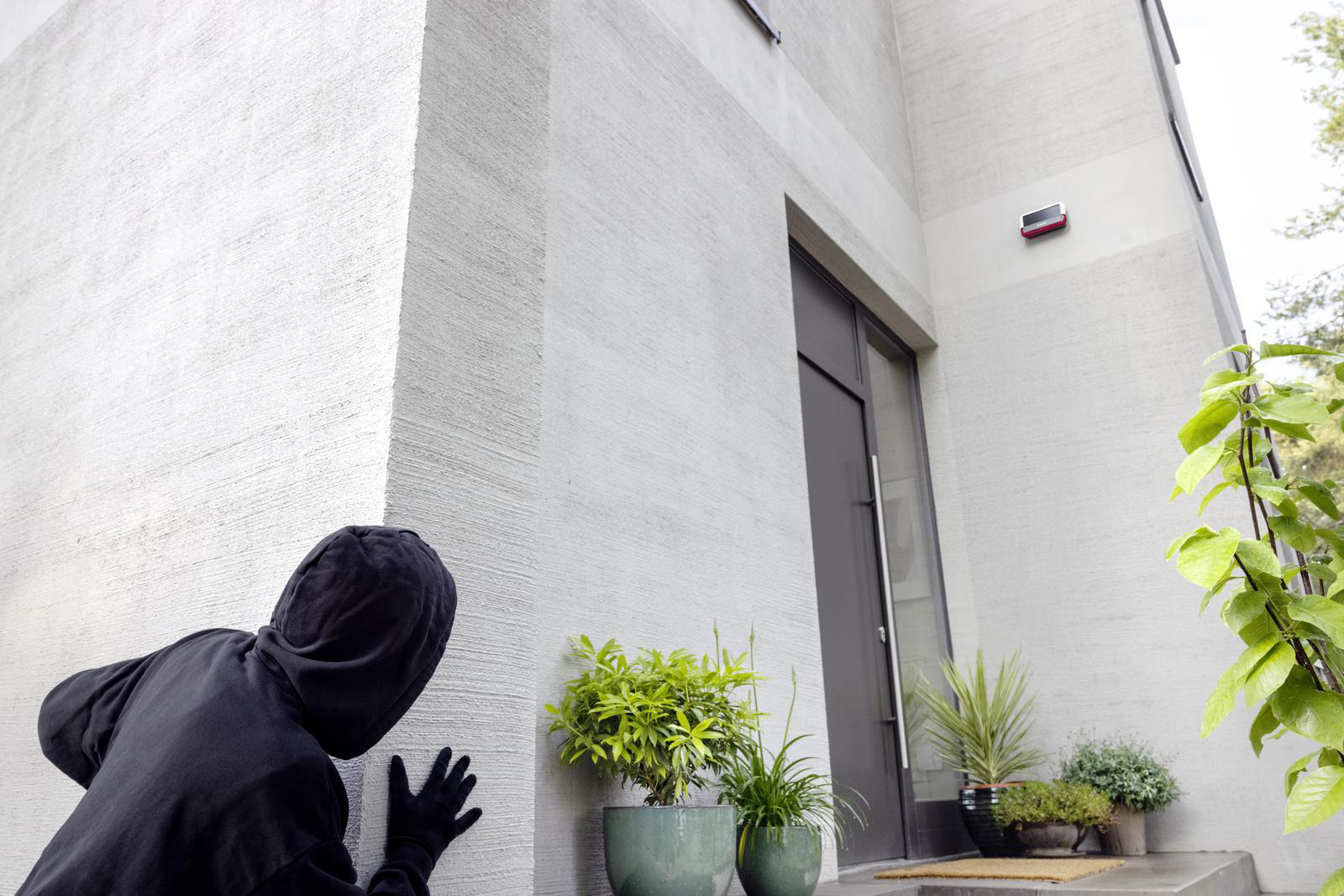Eine Person in schwarzer Kapuzenkleidung, die um eine graue Gebäudeecke späht.