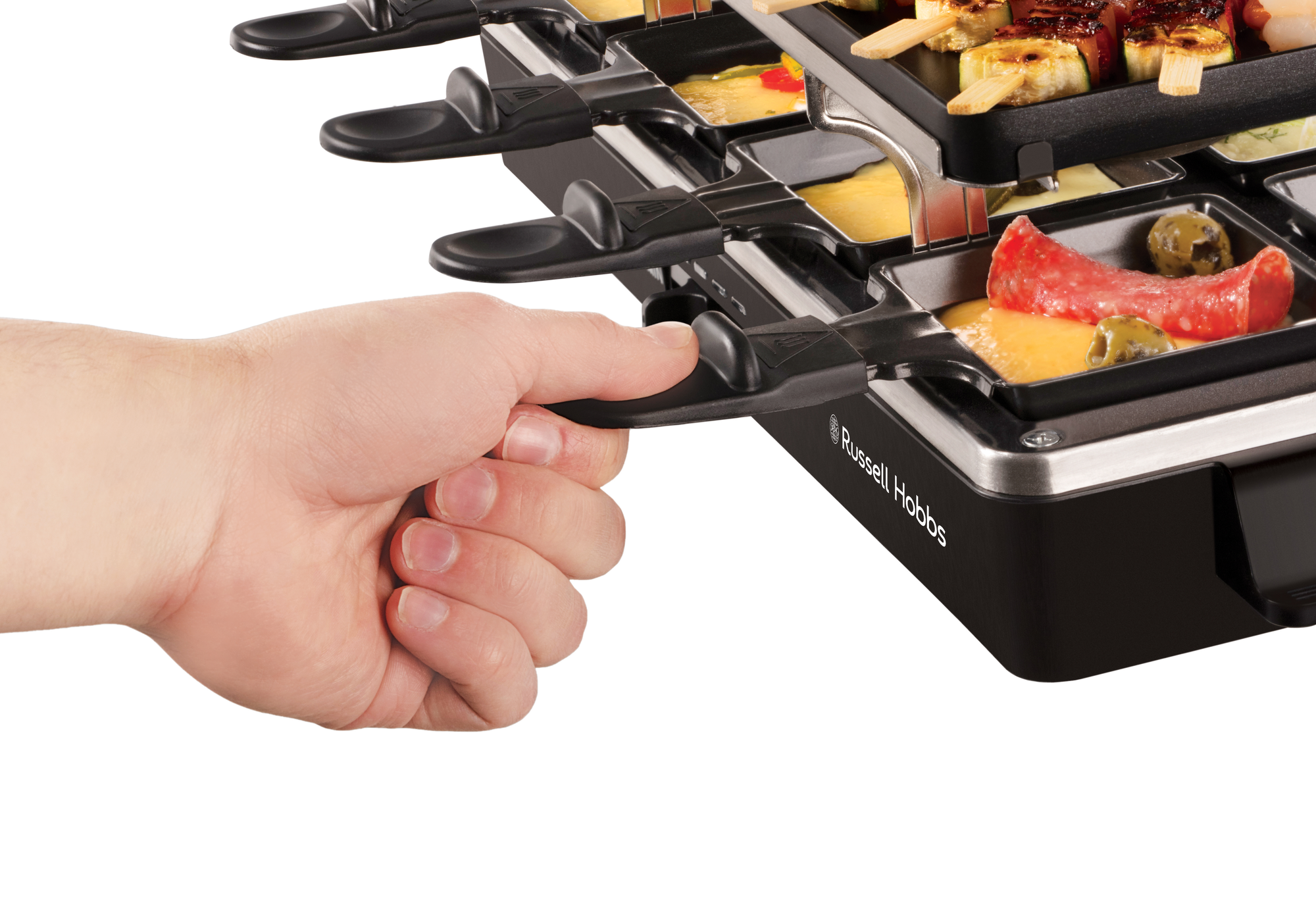 Egy kéz helyez egy tálcát étellel a raclette grillbe, sajt és egyéb ételek láthatók.