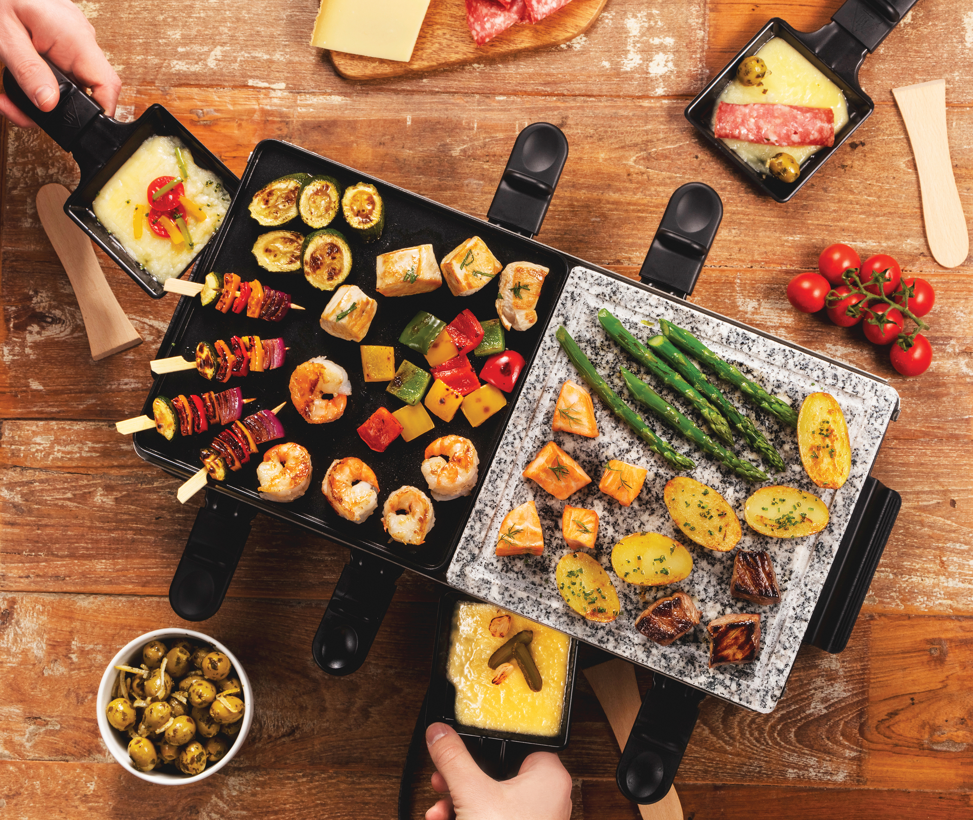 Raclette grill különféle főtt ételekkel, mint zöldségek, húsok és sajt, fa asztalon.