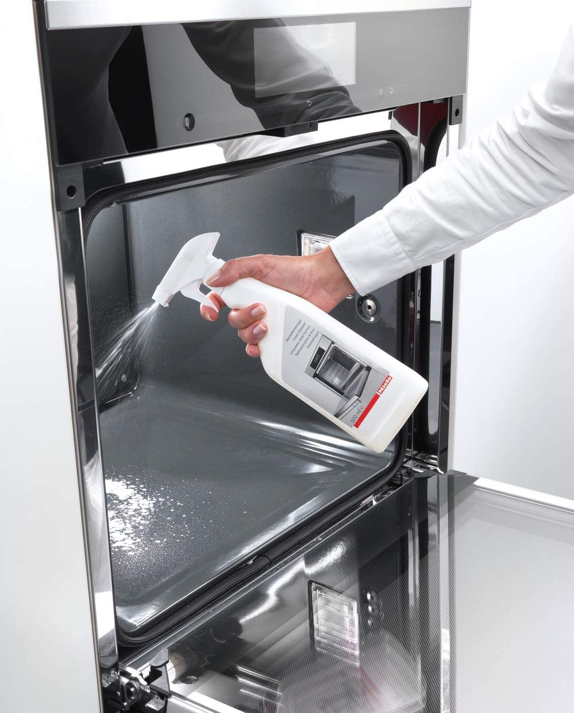 A person sprays cleaning solution inside an oven. A cleaning bottle is held in front of the open oven door.